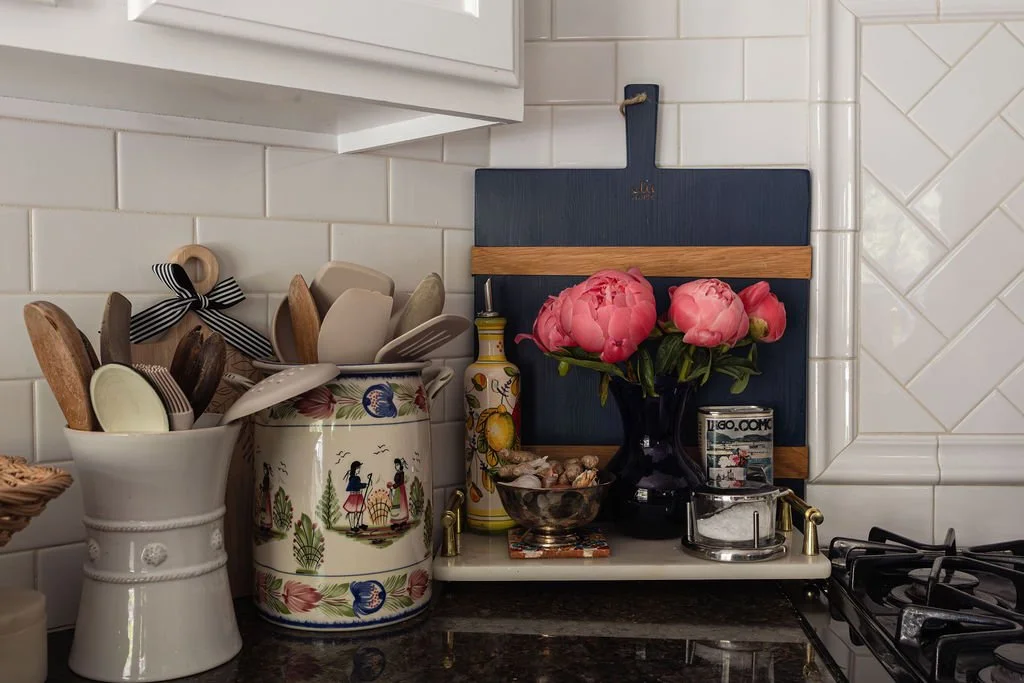 Kitchen countertop with utensils in ceramic containers, a small bowl of shells, a black vase with pink peonies, a decorative cutting board, and a spice jar.