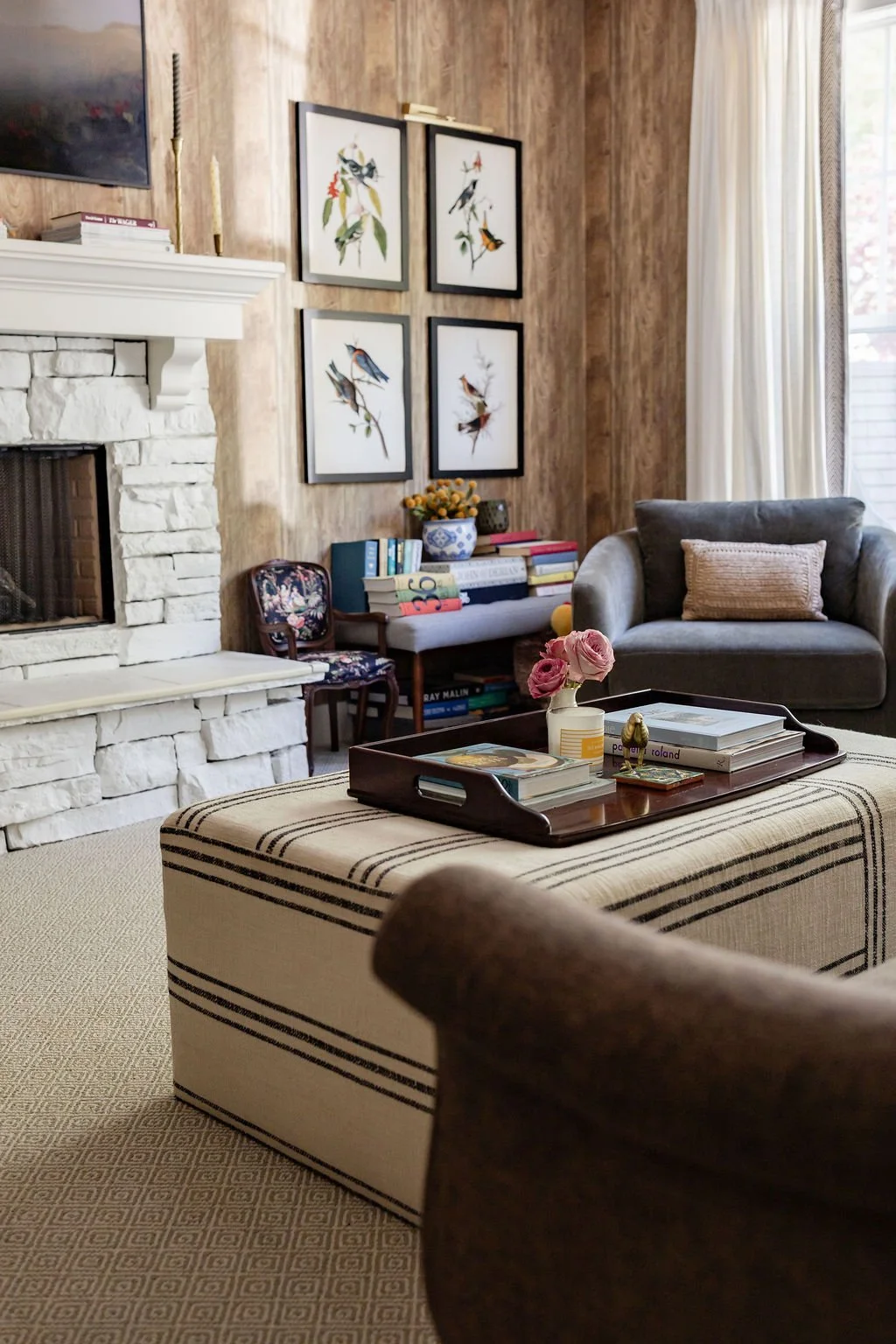 A cozy living room with a white stone fireplace, framed bird artwork on a wooden wall, a gray armchair, a floral armchair, and a bench with striped upholstery. There is a black tray with books and a pink flower in a white vase on the bench, a window 