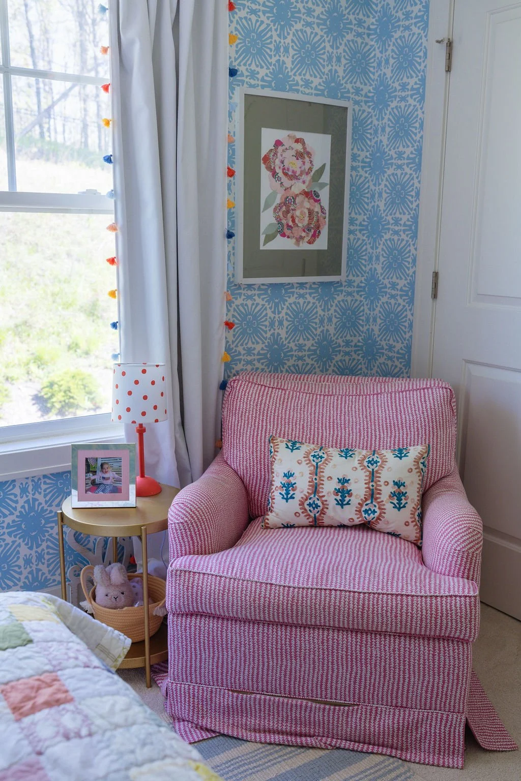A pink and white striped armchair with a cushion, beside a small round side table with a pink polka-dot lamp, a picture frame with a child’s photo, and a stuffed bunny in a basket, in a room with blue patterned wallpaper and a window with white curta