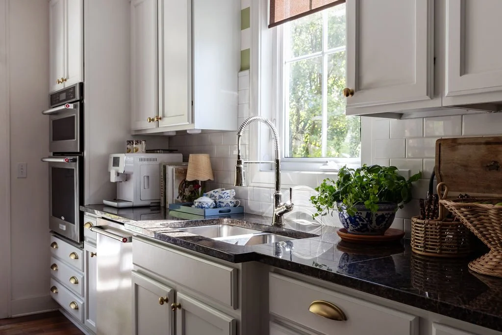 Kitchen with white cabinets, a black granite countertop, a window with green foliage outside, a plant in a decorative blue and white pot, a wicker basket, and kitchen appliances.