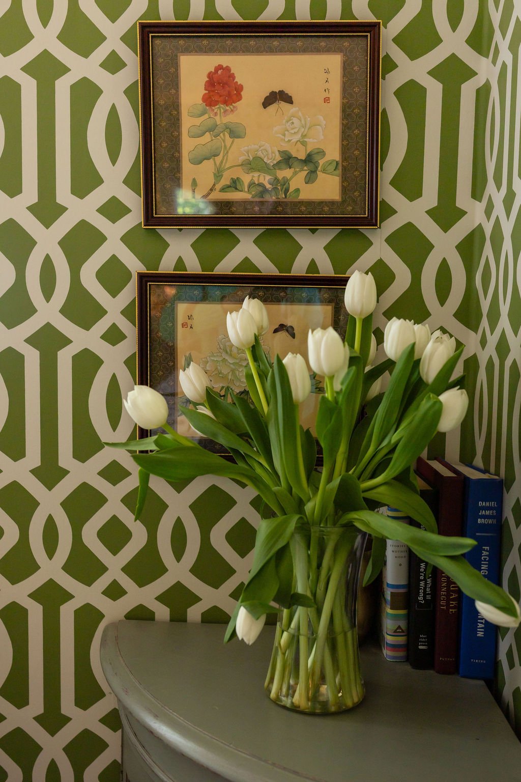 A vase of white tulips on a gray table in front of a green and white patterned wall, with two framed botanical prints and a row of books.