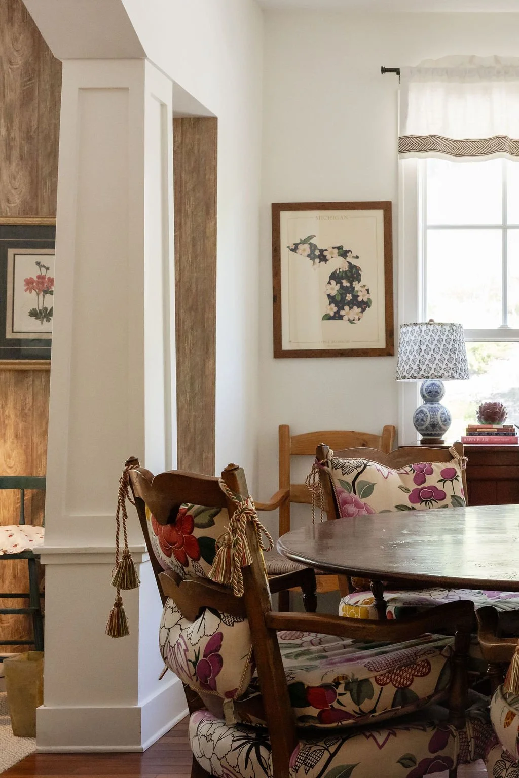 A cozy dining area with floral cushioned chairs around a wooden table, a window with curtains, a lamp with a patterned shade, and framed artwork on the wall.