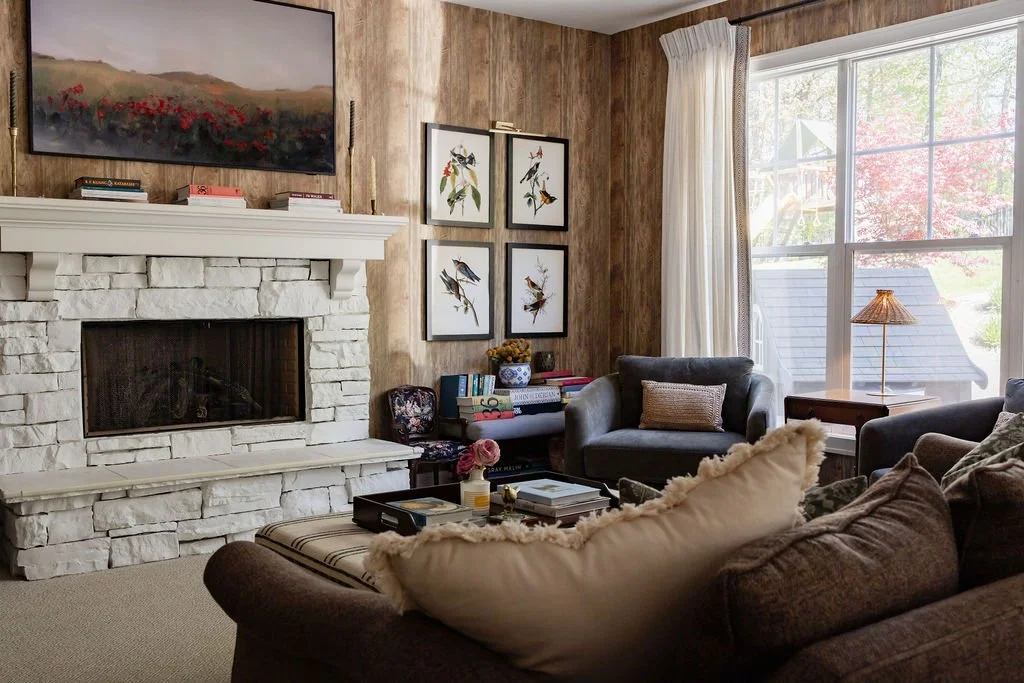 Cozy living room with a white stone fireplace, framed bird artwork, a large window with curtains revealing a backyard, and various couches and tables.