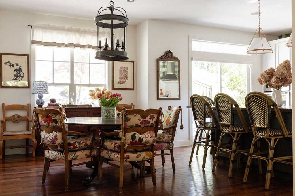 Bright dining area with a round wooden table surrounded by six floral upholstered chairs, a wooden sideboard with decorative items, a large window with sheer curtains, and a kitchen bar with six rattan chairs, decorated with vases of pink flowers.