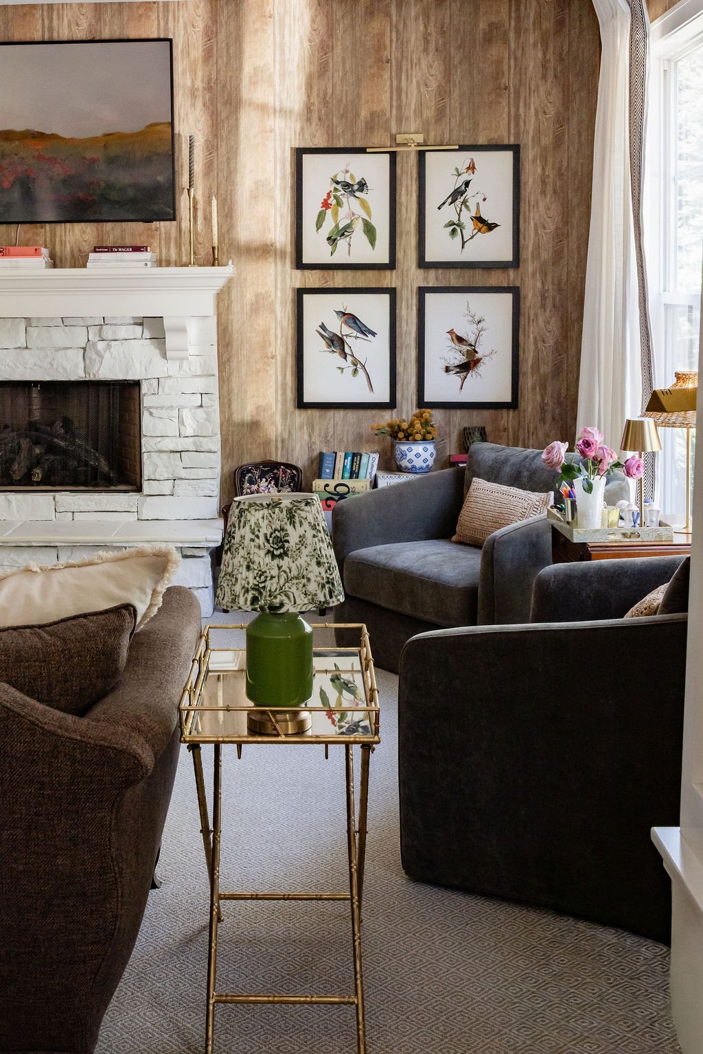 A cozy living room with a white brick fireplace, framed bird artwork on the wooden wall, a green table lamp, a gray armchair, and a side table with pink flowers, candles, and books near a window with white curtains.