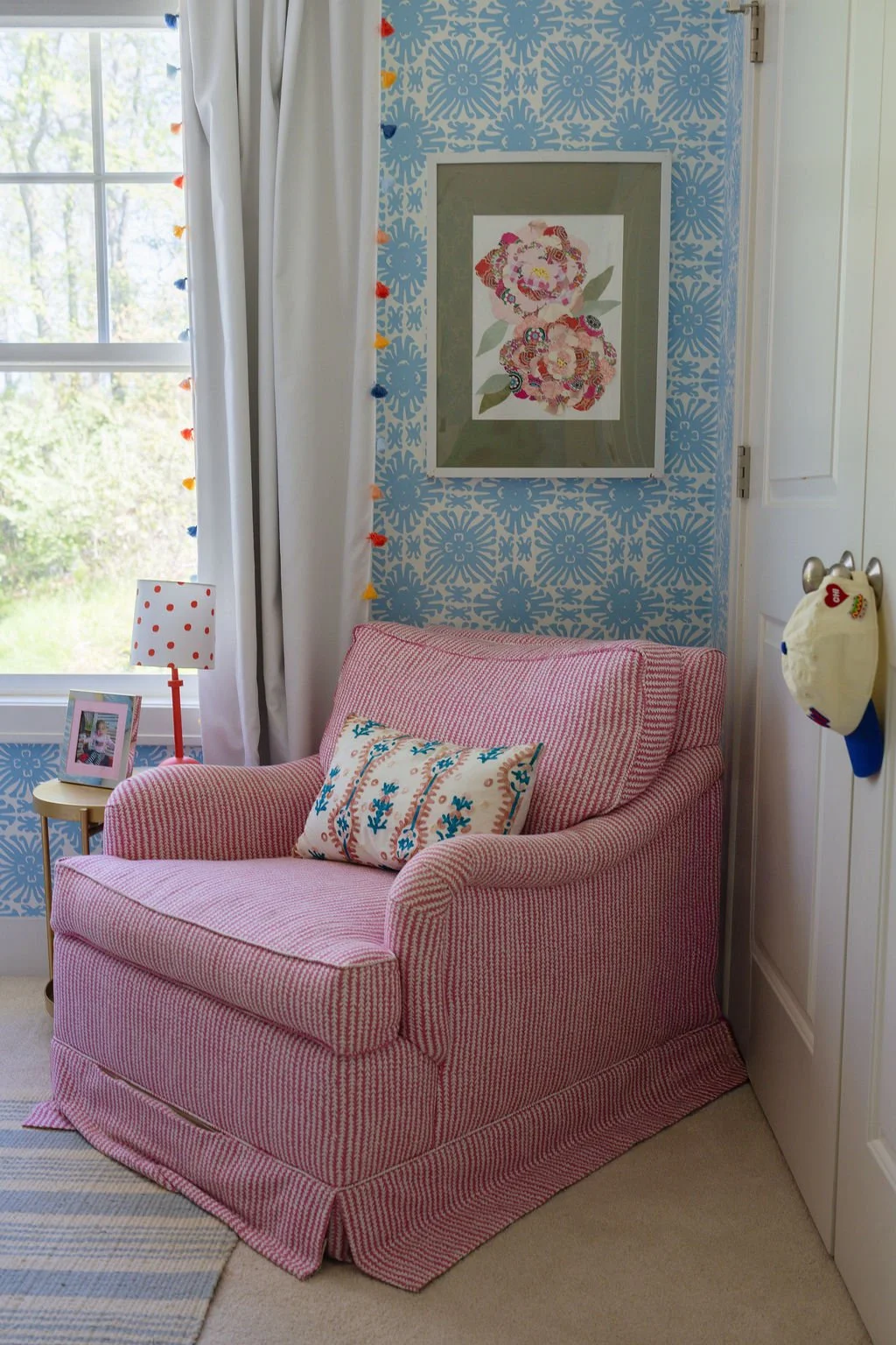 A cozy corner in a room with a pink upholstered armchair, a patterned pillow, a small side table with a framed photo and a pink and white polka-dot lamp, a window with white curtains, a floral wall art, and a wall with blue patterned wallpaper.
