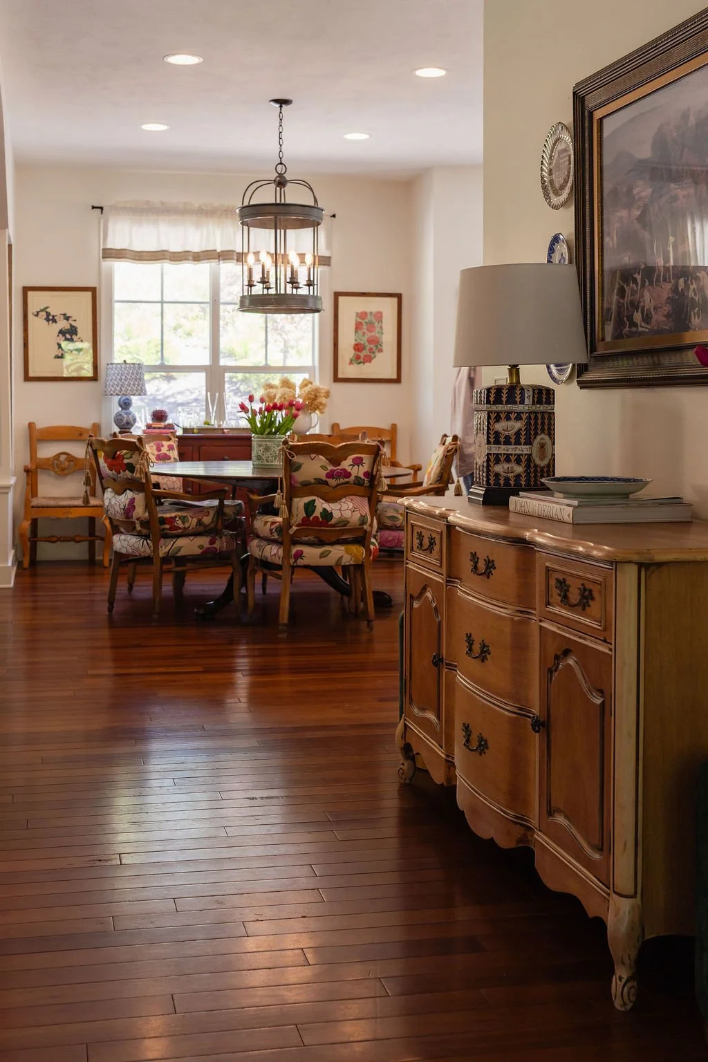 A cozy dining room with a wooden dining table surrounded by floral upholstered chairs, a round window with white curtains, a chandelier hanging from the ceiling, and vintage decor including a sideboard with a lamp, books, and plates.