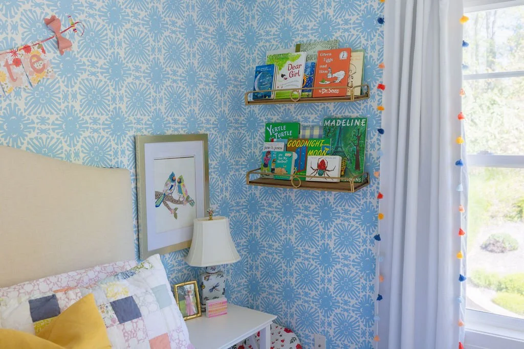 A bedroom corner with a bed, a lamp, framed art, and books on wall-mounted shelves. The room has blue floral wallpaper, a window with curtains, and colorful pom-pom string lights.