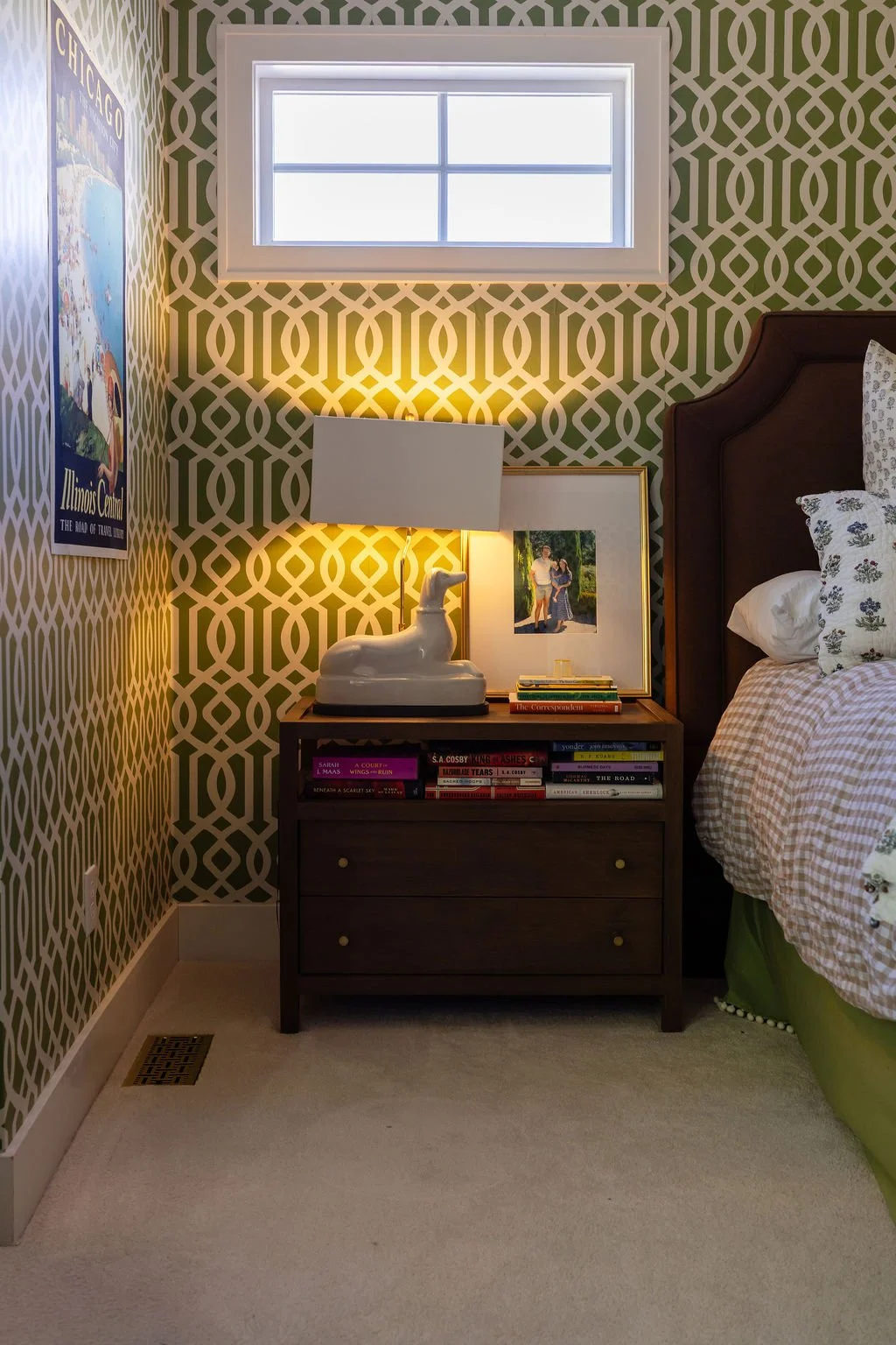 A bedroom corner with geometric patterned wallpaper, a small window, a wooden nightstand with a large ceramic dog lamp, framed artwork, and a stack of books. Part of a bed with floral and gingham bedding is visible.