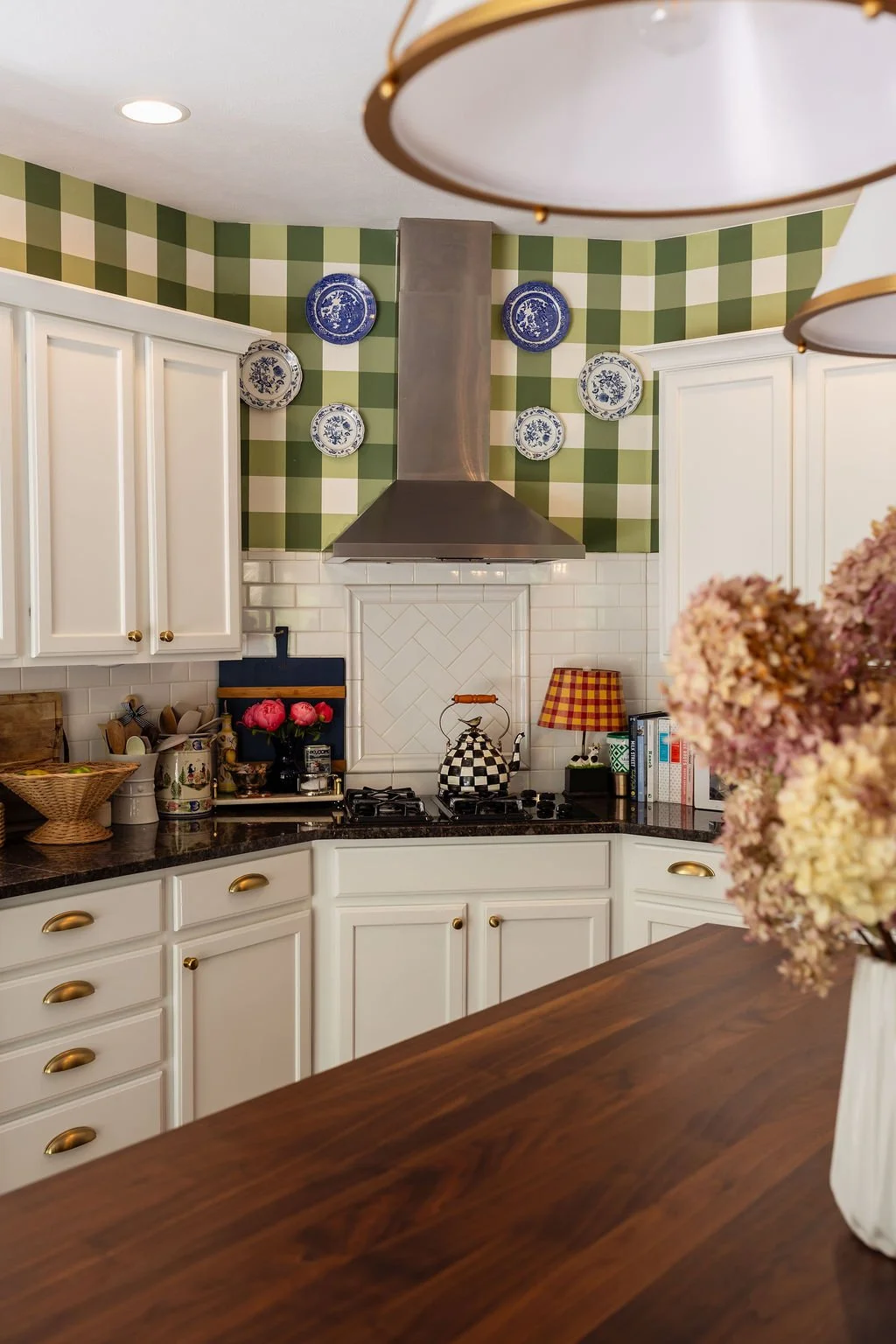 A cozy kitchen with white cabinets, black countertops, a stainless steel range hood, and decorative blue and white plates on a green and white checkered wall. The countertop features a checkered teapot, a small lamp with a yellow and red checkered sh