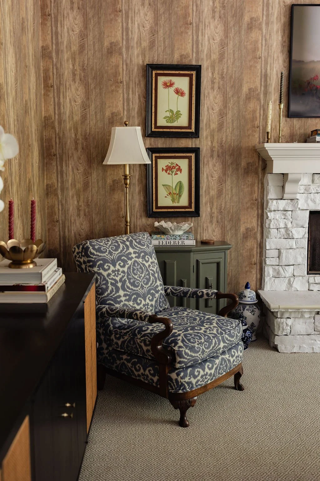 A cozy living room corner featuring an armchair with patterned upholstery, a green side cabinet with books and decorative items, two framed floral prints on a wood-paneled wall, a floor lamp, and a white brick fireplace with candles on the mantle.