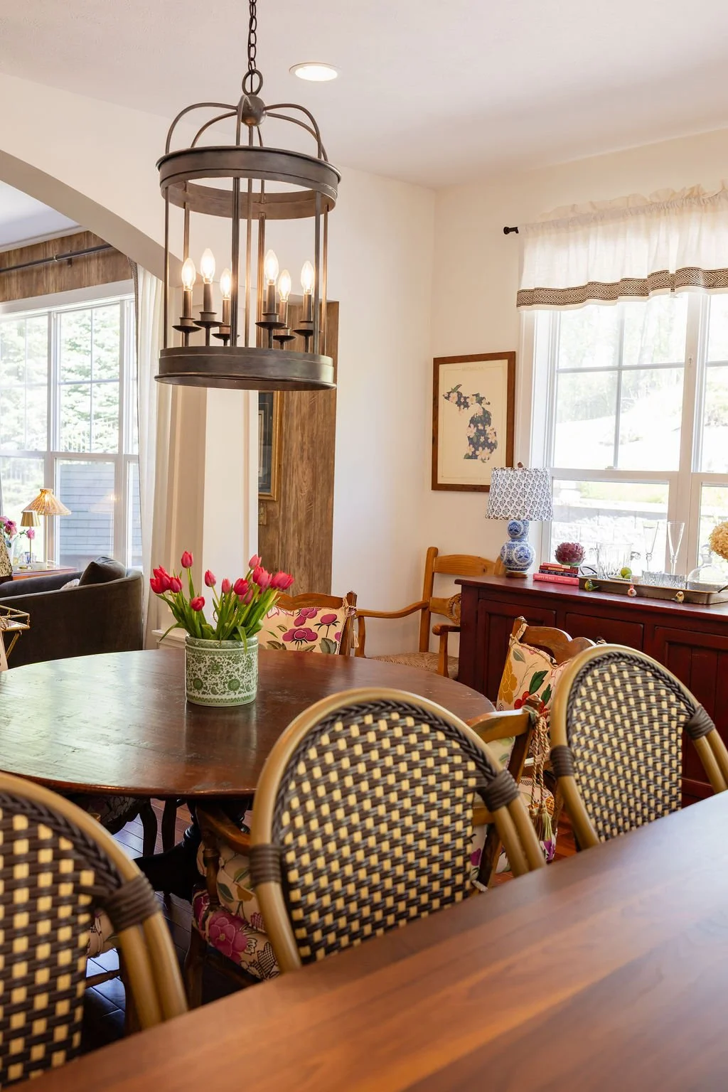 A dining room with a round wooden table, pink tulips in a green vase, woven chairs, a dark wood sideboard with books, a blue and white lamp, and large windows letting in natural light.