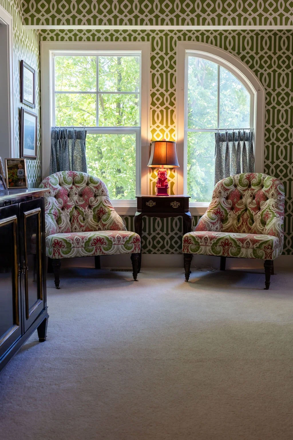Living room with two patterned armchairs, a small table with a lamp between them, window with tree view behind, patterned wallpaper, and a sideboard partially visible on the left.
