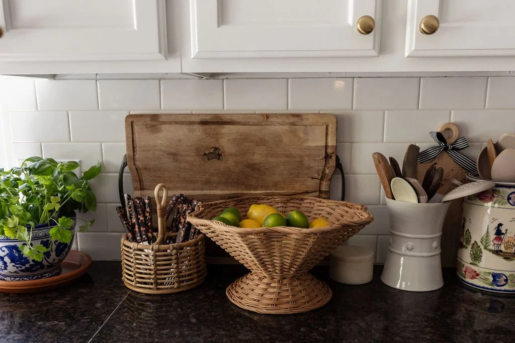 Kitchen counter with a potted basil plant, wicker utensil holders, a wooden cutting board, a basket of lemons and limes, and various dishware in containers.
