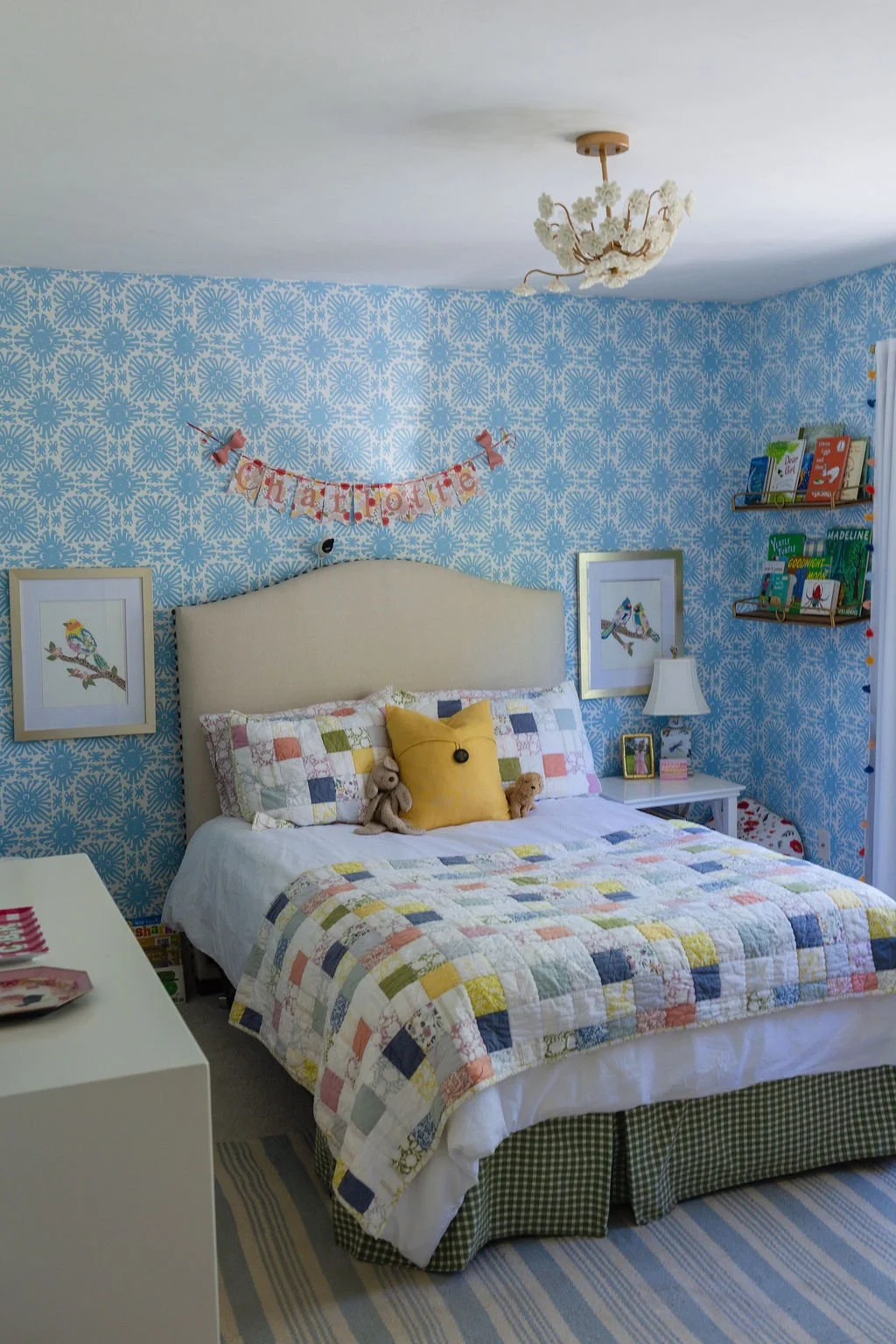 A cozy bedroom with a bed covered by a multicolored quilt, a yellow pillow, and plush toys. The blue patterned wallpaper features framed bird artwork. A chandelier hangs from the ceiling, and a "happy birthday" banner is displayed above the bed. A white nightstand with a lamp and picture frame sits beside the bed. Books are on a wall-mounted shelf, and a white dresser is partially visible.