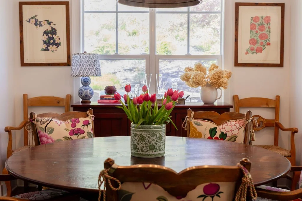A round wooden dining table with four chairs, one at each side. The chairs have floral-patterned cushions tied to the backrests. A potted plant with pink tulips is in the center of the table. Behind the table, a sideboard with a blue and white lamp, 
