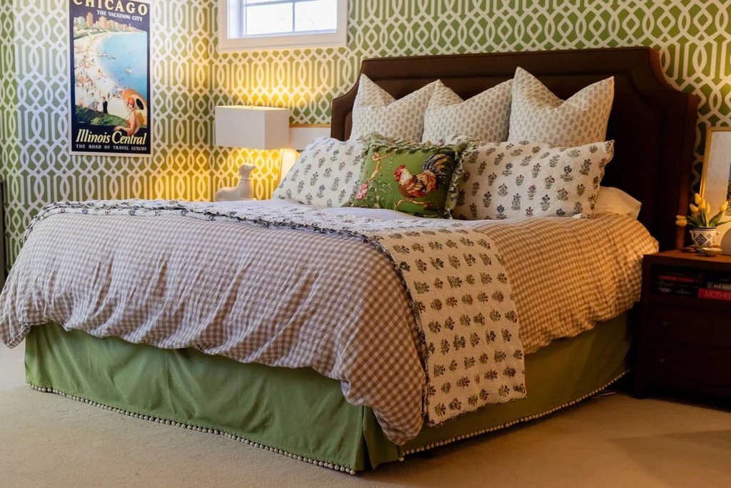A neatly made bed with a green bed skirt, patterned bedding, and multiple pillows, in a bedroom with green and white patterned wallpaper, a nightstand with a potted plant, and a decorative poster on the wall.