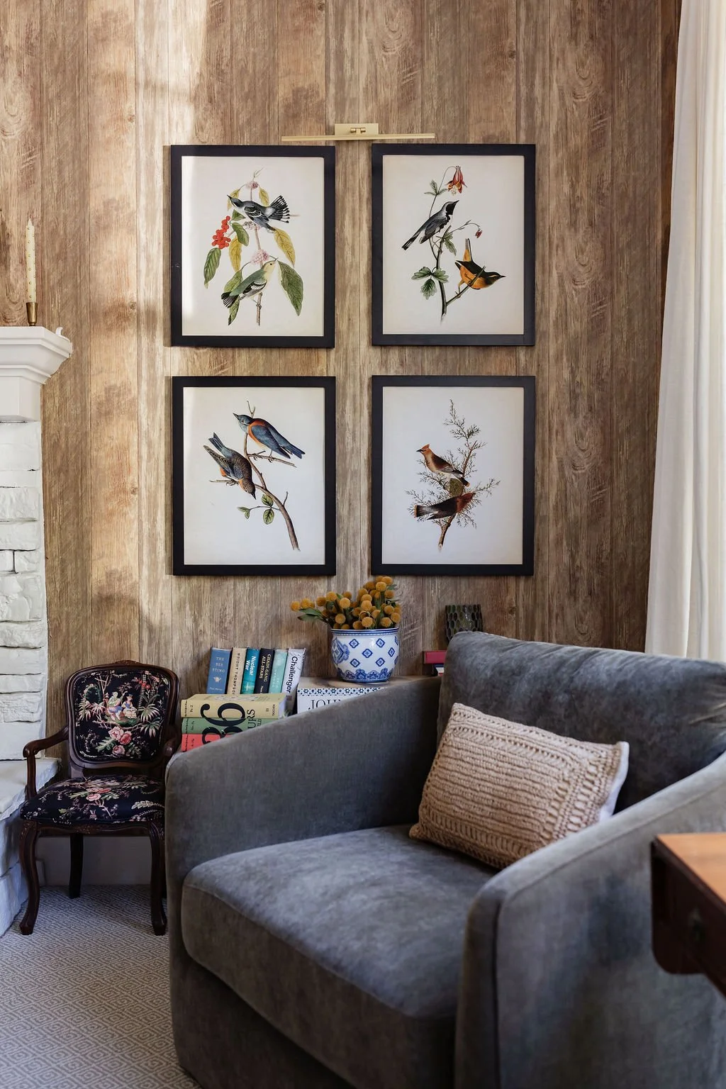 A living room corner with four framed bird illustrations on a wooden wall, a gray sofa with a beige pillow, a floral upholstered chair, a shelf with books and a vase with yellow flowers, and a white curtain on the right.