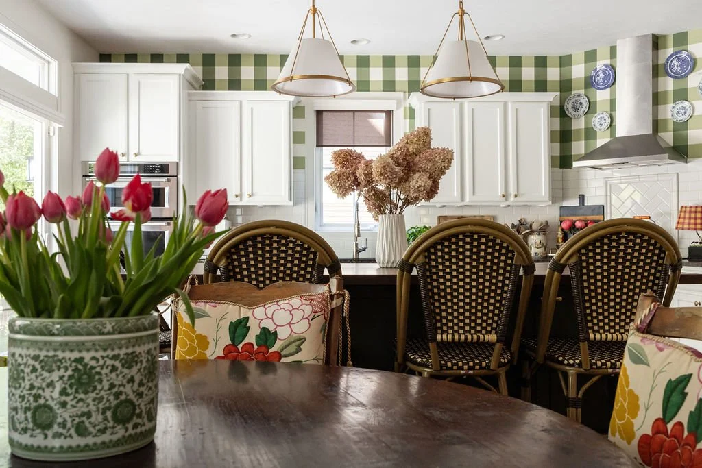 A bright kitchen with white cabinets, green checkered wallpaper, and a dining area with floral chairs. A vase with pink dried flowers sits on the black kitchen island, and pink tulips are in a decorative pot on a wooden table.