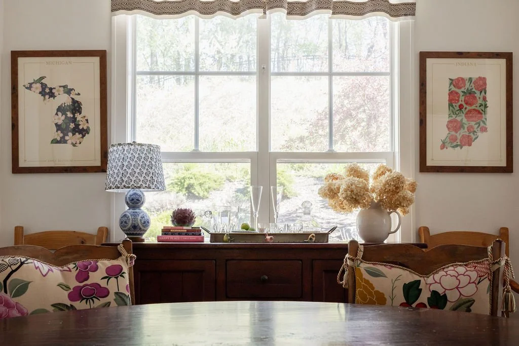 Dining room with a wooden table, floral pillows, a windowsill with a vase of white hydrangeas, a lamp with a patterned shade, framed floral artwork of Michigan and Indiana, and trees visible through the window.