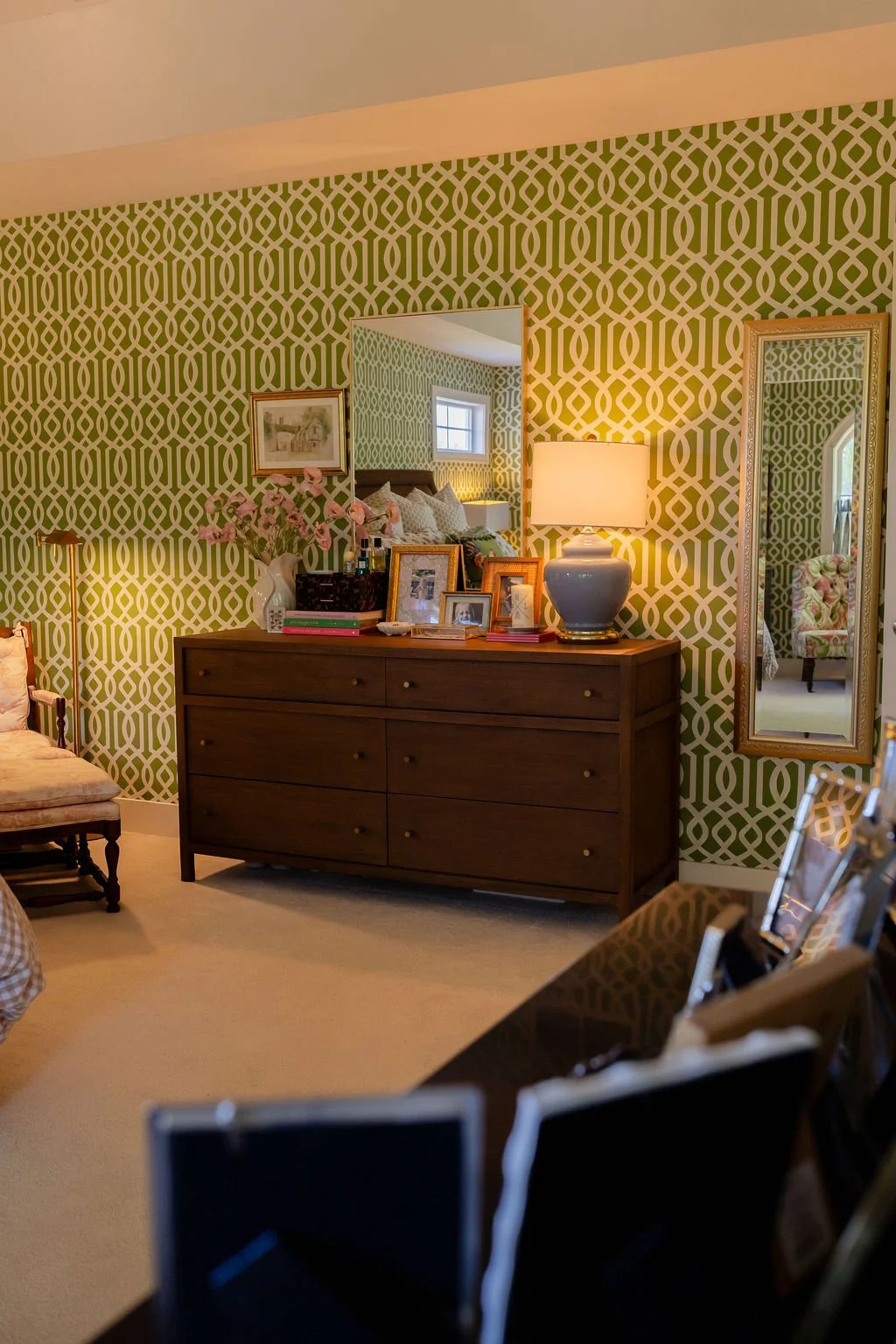 A bedroom with green patterned wallpaper, a wooden dresser topped with framed photos, a vase with pink flowers, a lamp, and a mirror reflecting part of the room.