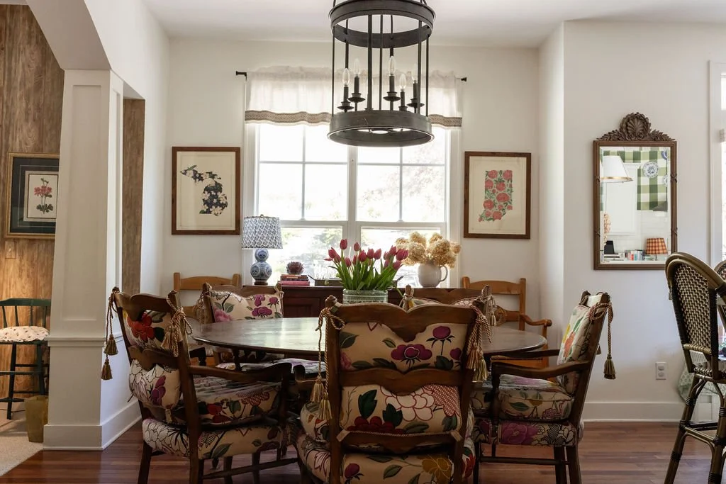 A cozy dining room with a round wooden table, floral cushioned chairs, and a window with white curtains. Decor includes framed floral art, a chandelier, a lamp with a patterned shade, and vases with flowers.