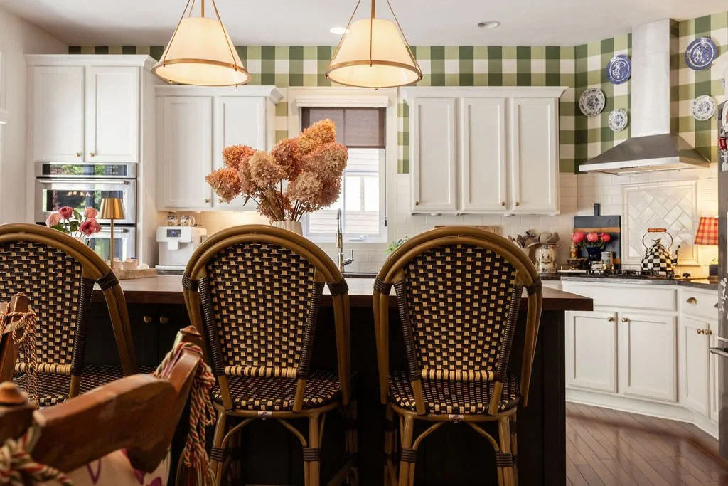 A cozy kitchen with white cabinets, a green plaid backsplash, and hanging pendant lights. There is a large vase with pink flowers on the counter, and decorative plates on the wall. The kitchen has a stove, oven, and a window behind the sink.