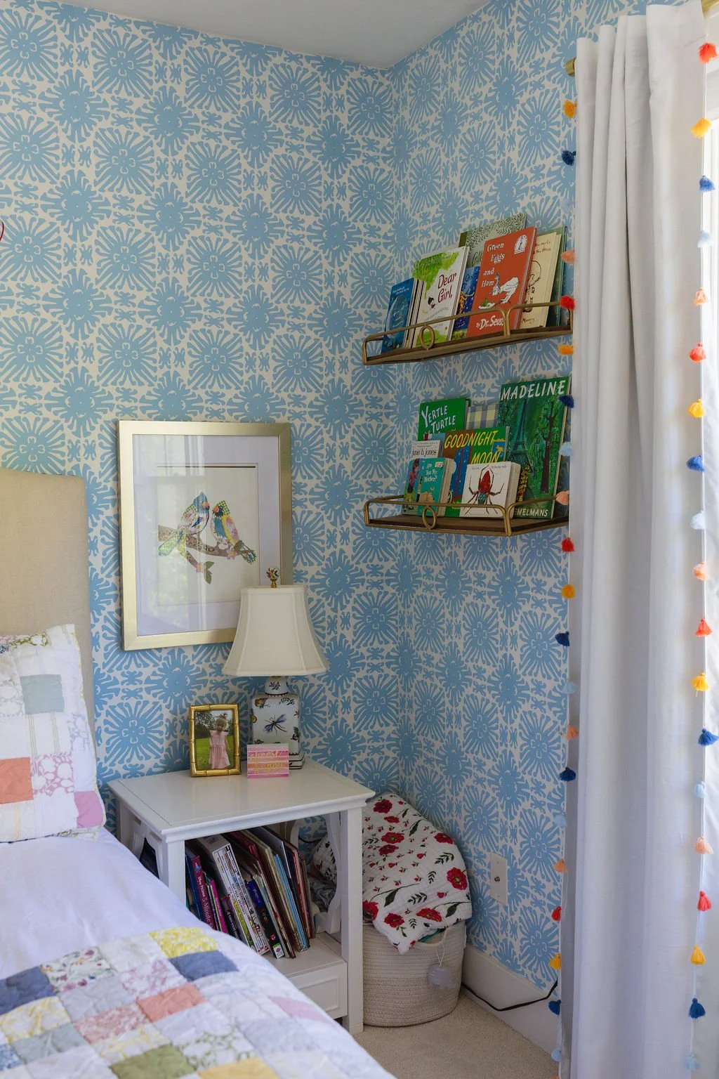 A cozy bedroom corner with a blue patterned wallpaper, a white bedside table with books and framed photos, a table lamp, a framed bird artwork, and colorful books on wall-mounted shelves. White curtains with multicolored pom-pom trim hang beside a wi
