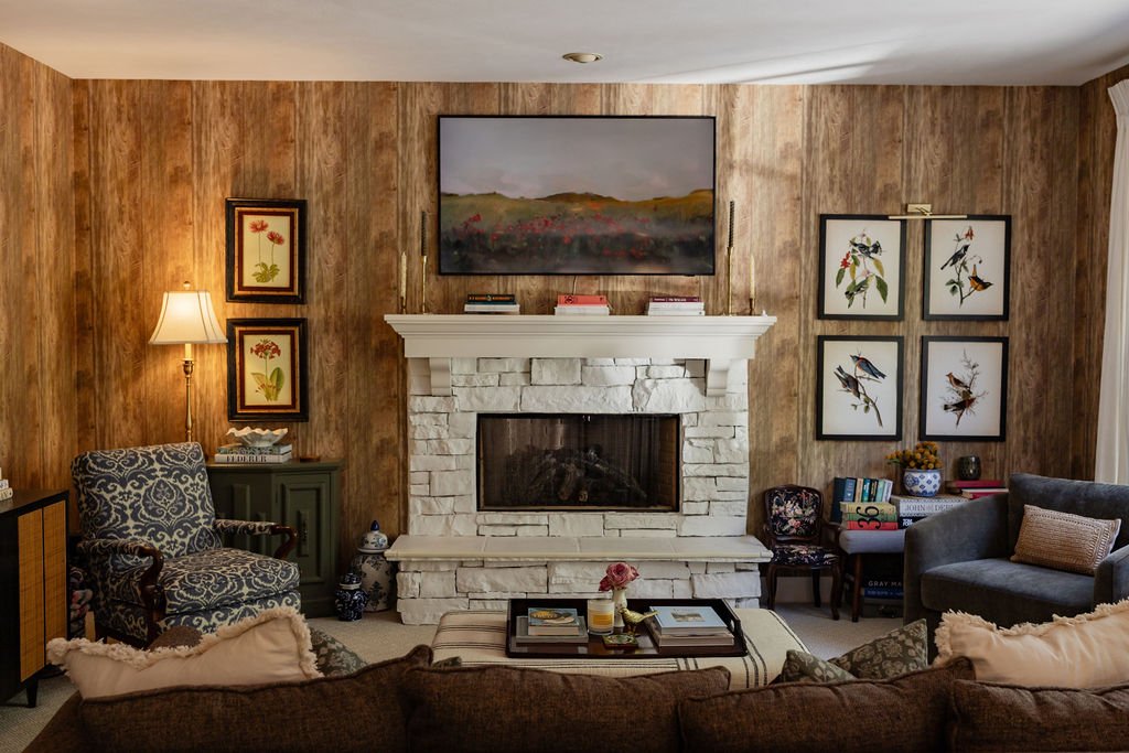 Living room with wood-paneled walls, white stone fireplace, wall art featuring birds and flowers, armchair, side table with lamp, and a sofa with cushions.