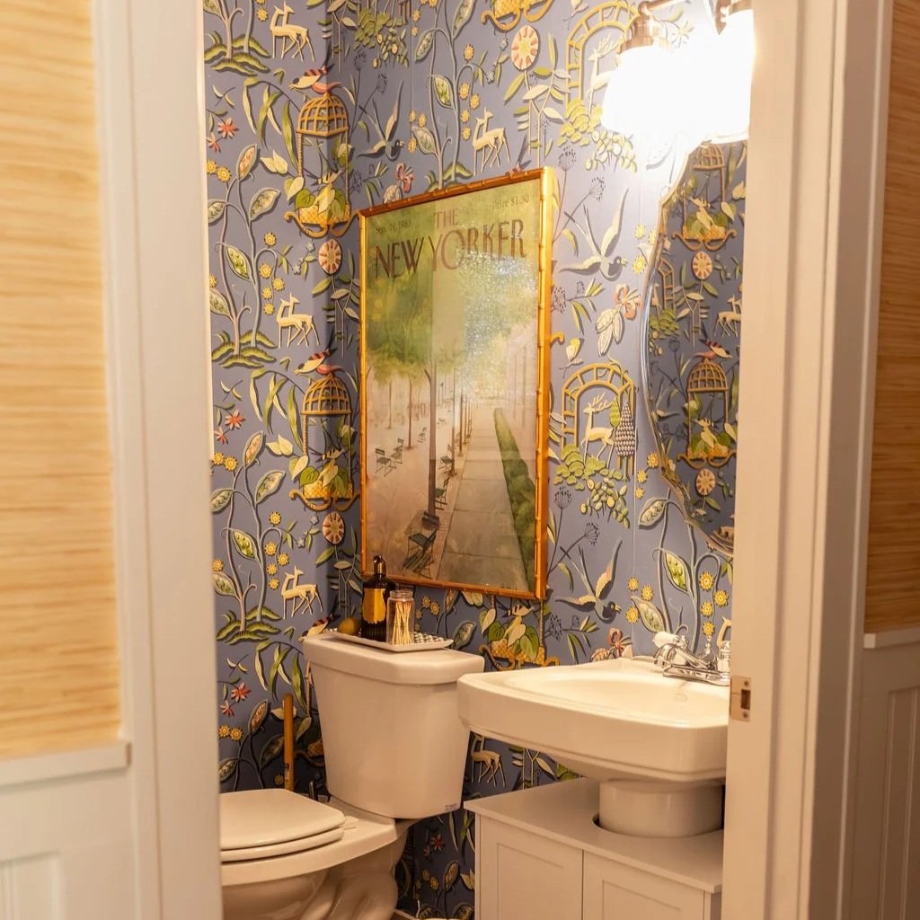 A small bathroom with blue wallpaper featuring a floral and bird pattern, a mirror with the word “The New Yorker” on it, a white toilet, a white sink with a cabinet, a soap dispenser, and a wall-mounted light fixture.
