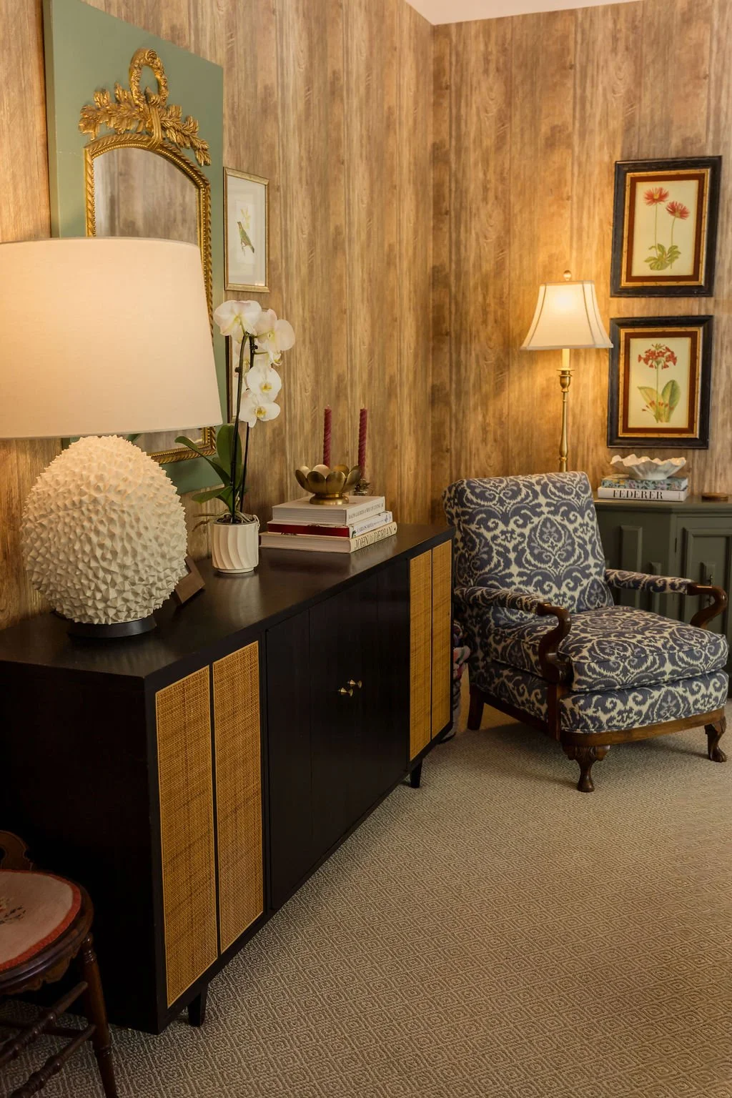 A cozy living room corner with a dark wood cabinet topped with a white textured lamp, a white orchid in a pot, stacked books, and red candles. An armchair with blue and white patterned upholstery sits next to a green sideboard, with framed botanical 
