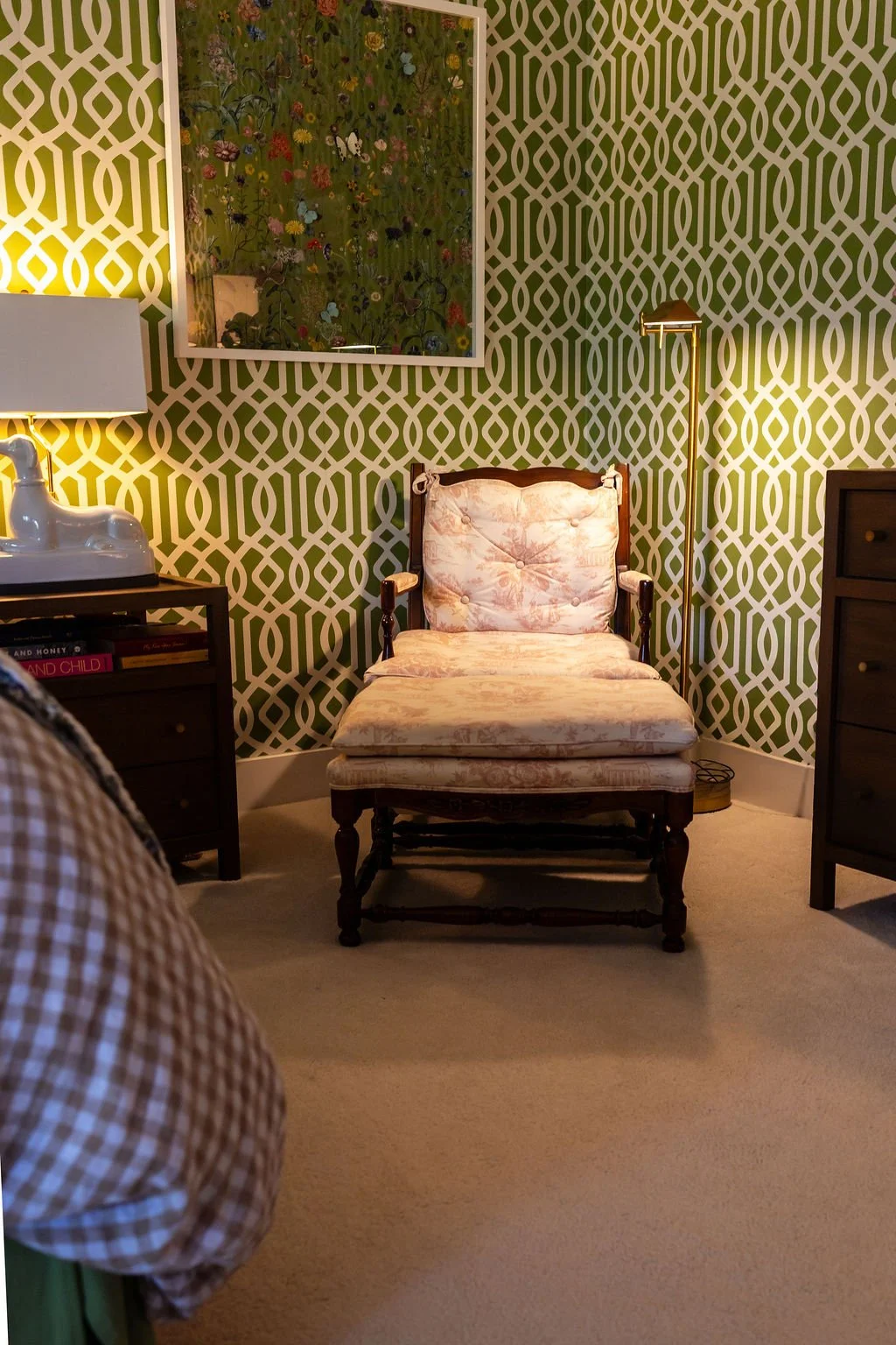 Comfortable vintage armchair with floral upholstery in a cozy room with patterned wallpaper, a small table with a lamp, and framed artwork on the wall.