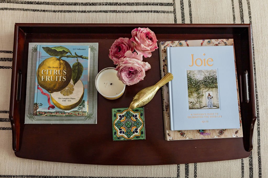A wooden tray holding a book titled 'The Book of Citrus Fruits', a book titled 'Joie', a small bouquet of pink roses, a lit white candle in a glass container, a gold fish-shaped item, and a colorful ceramic tile, all placed on a beige fabric surface 