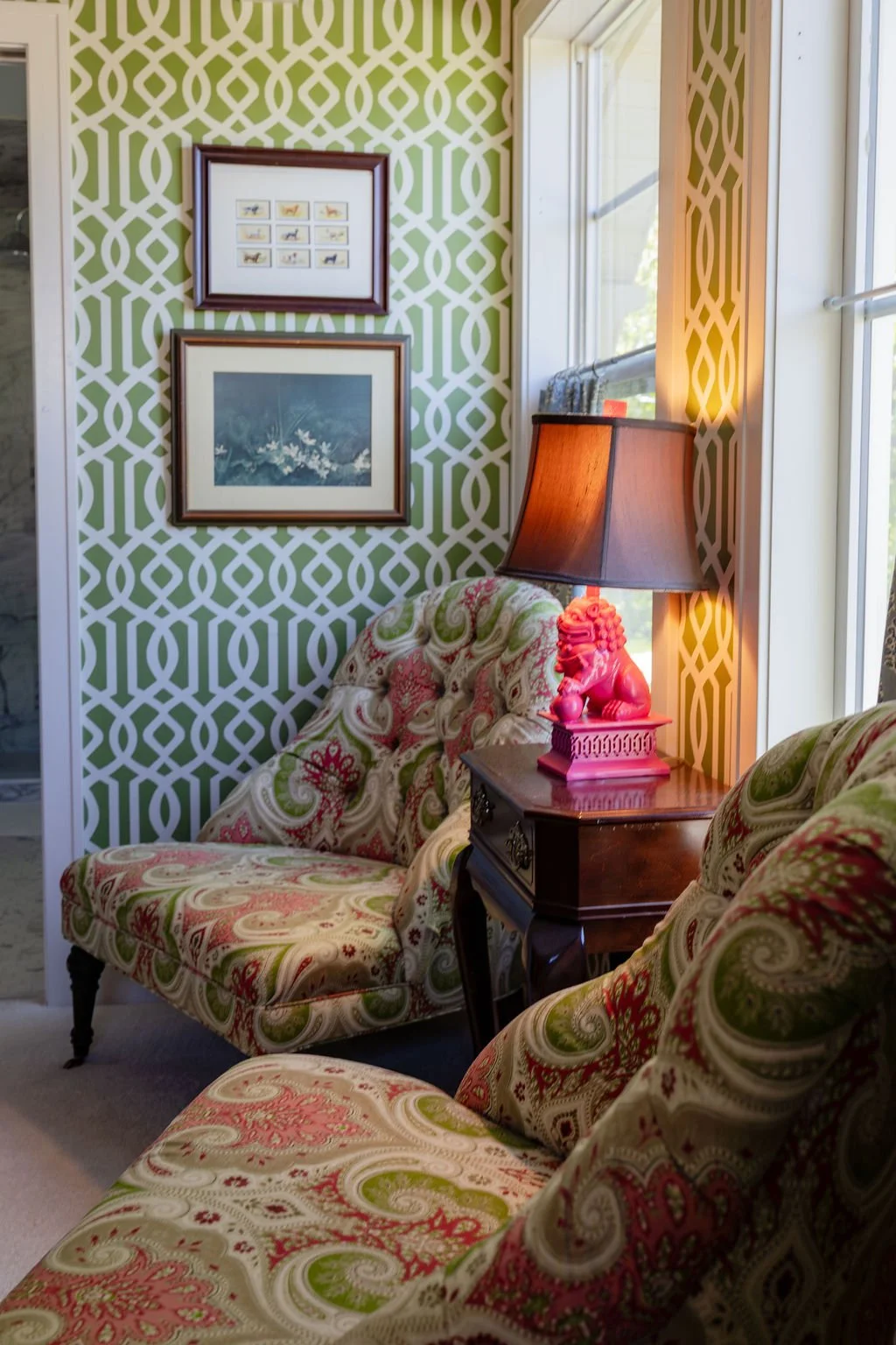 A cozy living room corner with two patterned armchairs, a side table with a pink lamp featuring a lion statue, and two framed pictures on a green and white geometric wallpaper.