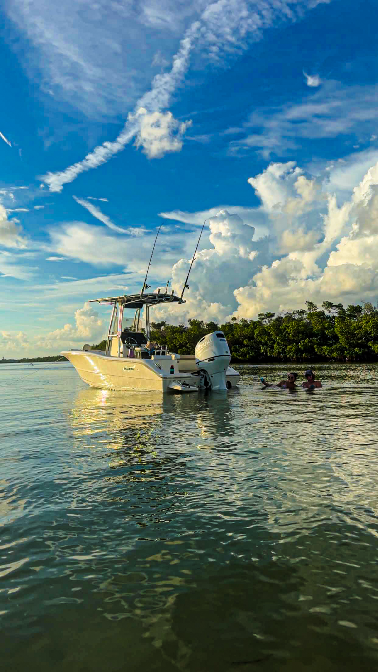 A boat floating on a body of water with two people swimming nearby, a cloudy sky above, and trees in the background.