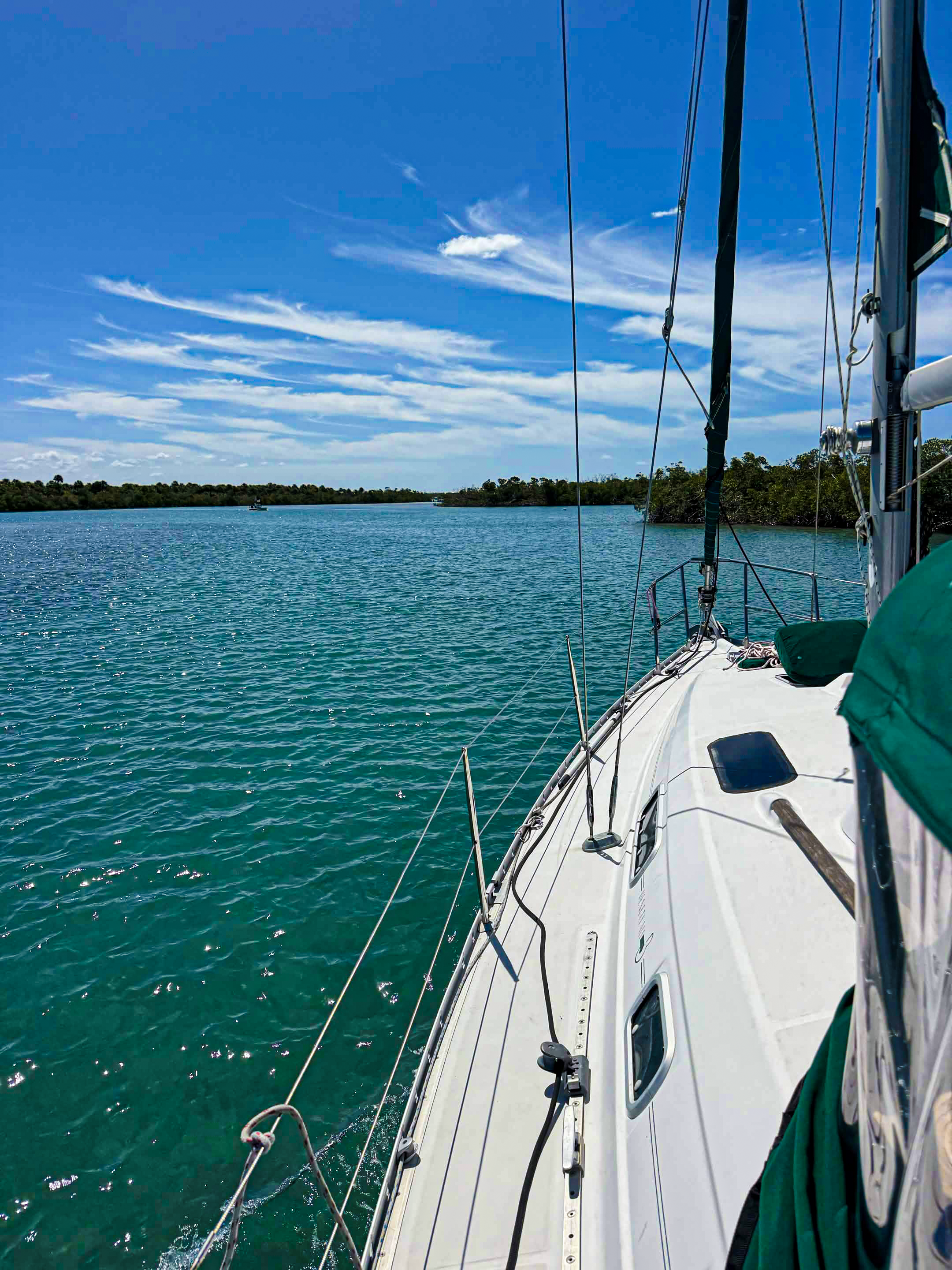 View from a sailboat on clear, blue water with distant boat and green shoreline under a partly cloudy sky.