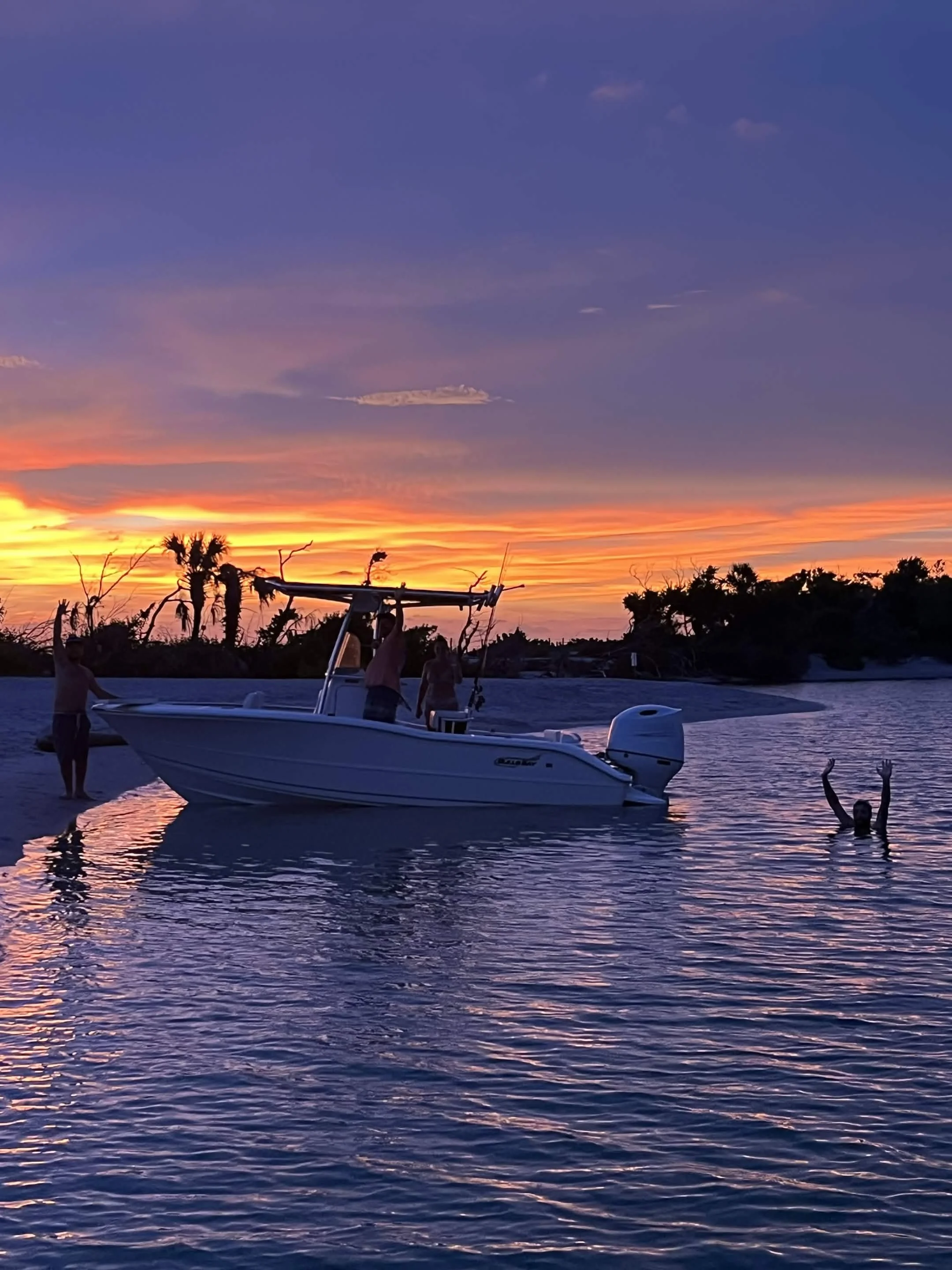 A boat floating on calm water during a colorful sunset with clouds in the sky. Several people are in and around the boat, with some standing and others in the water, enjoying the scene.