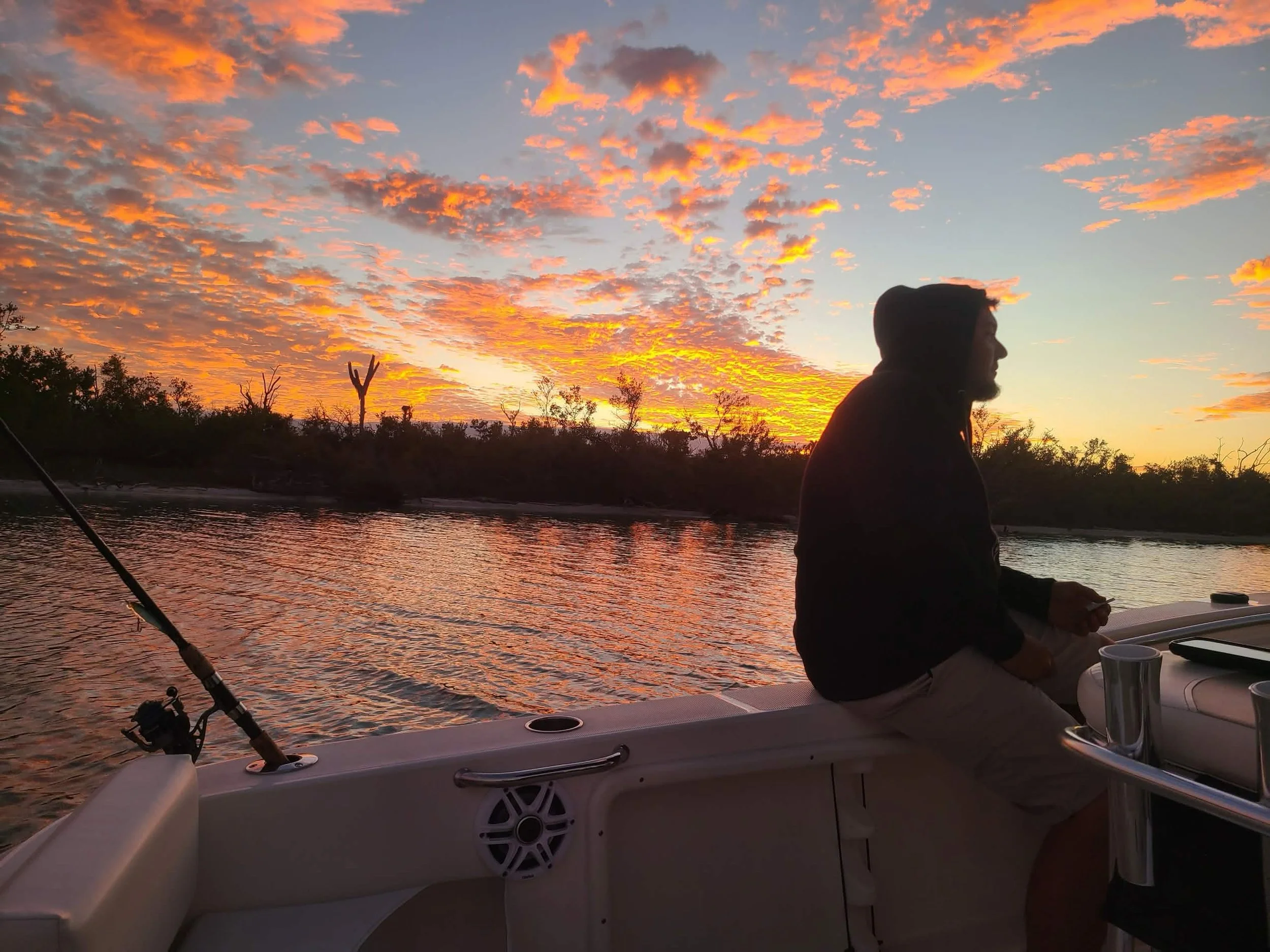 A person sitting on a boat during sunset, fishing by the water, with a sky full of orange and pink clouds and trees in the background.