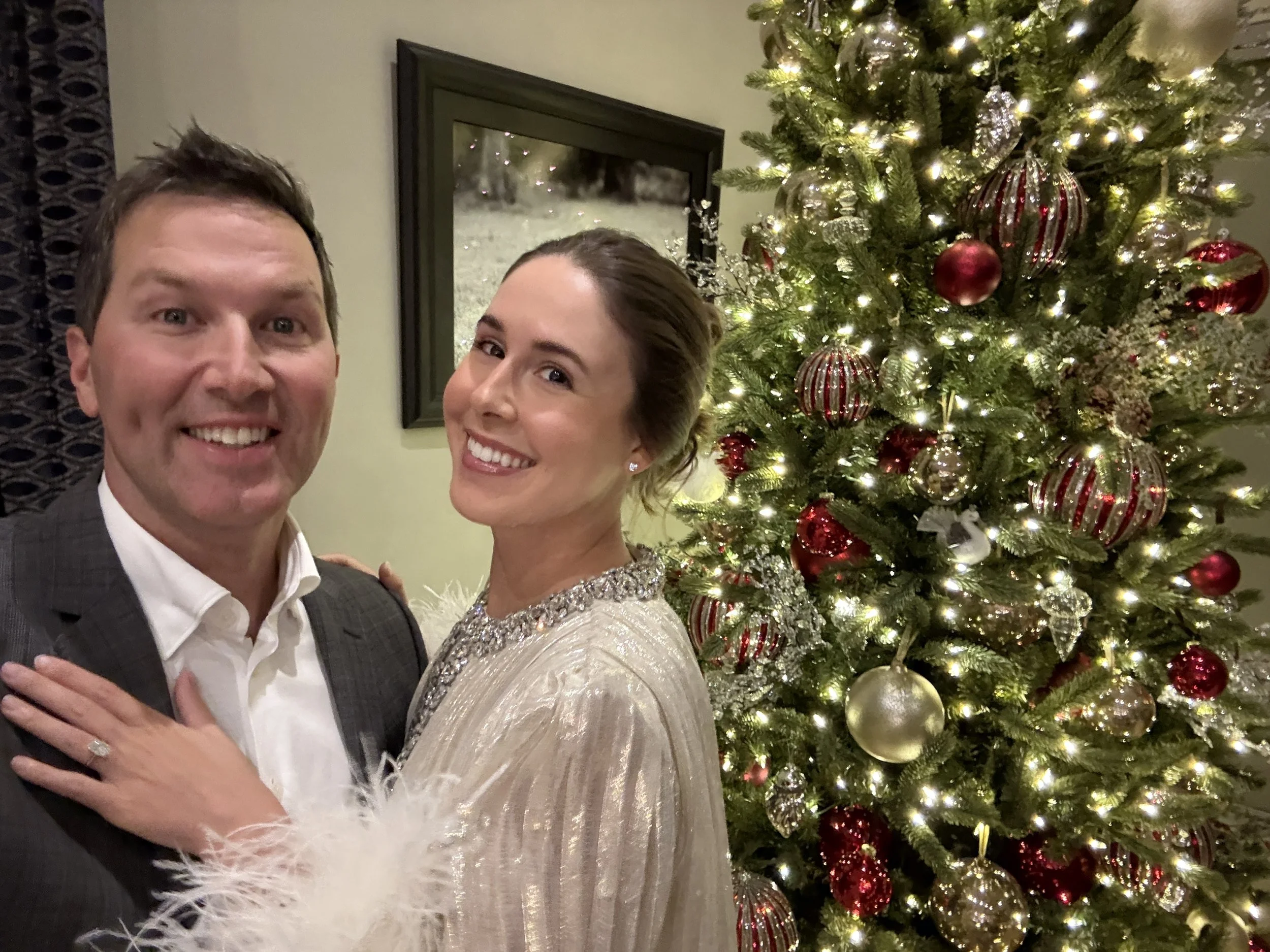 A man and woman smiling and embracing in front of a decorated Christmas tree with lights and red, gold, and silver ornaments.