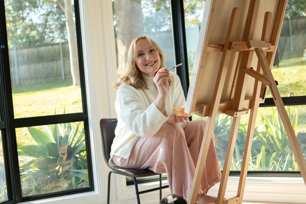 A woman sitting on a chair, smiling, holding a paintbrush and a palette while painting on a canvas set on an easel by large windows with a view of a green yard.