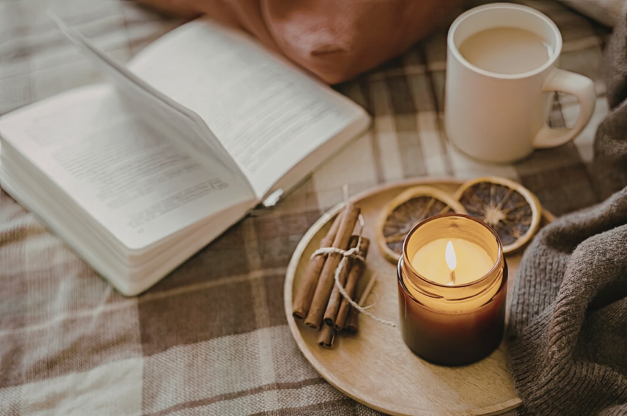 A placemat of a blanket with an open book, a coffee and a wooden plate with cinnamon sticks, dried orange slices, and a lit candle | Northwest Wellness | Therapy & Consulting in Thunder Bay