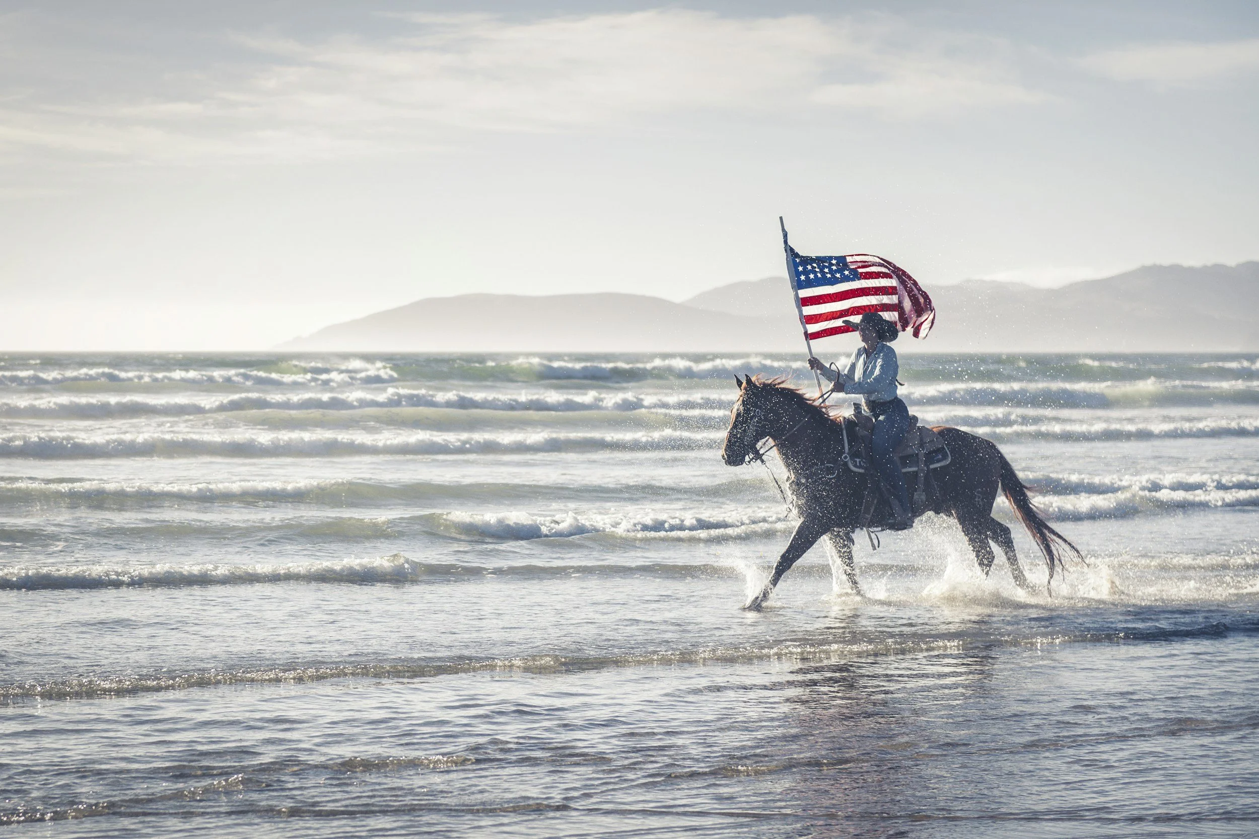 A person riding a horse in the ocean waves while holding an American flag, with a distant island and a partly cloudy sky in the background.
