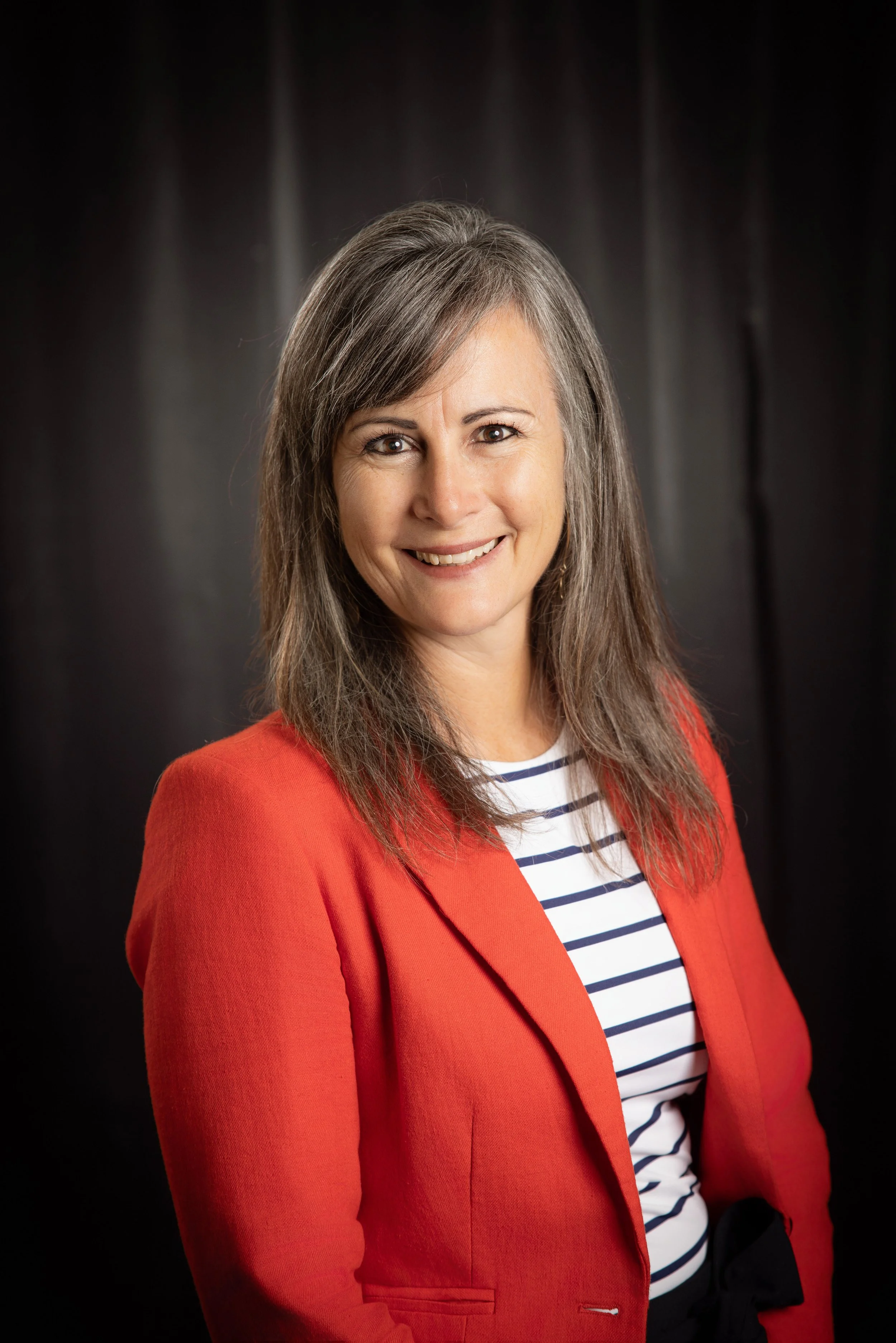 A woman with brown hair and a smile, wearing a red blazer over a white and navy striped shirt, against a dark background.