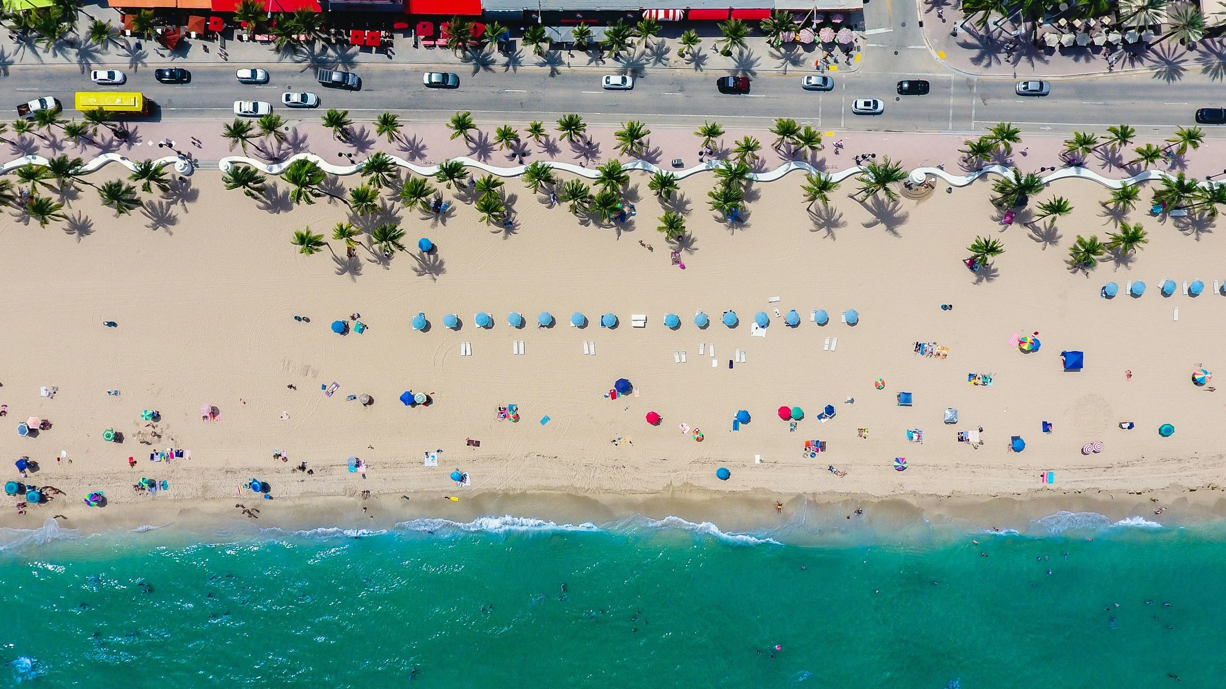 An aerial view of a beach with colorful umbrellas and people relaxing on the sand, a row of palm trees along the sidewalk, and a road with traffic running parallel to the beach. The ocean is visible at the bottom of the image.