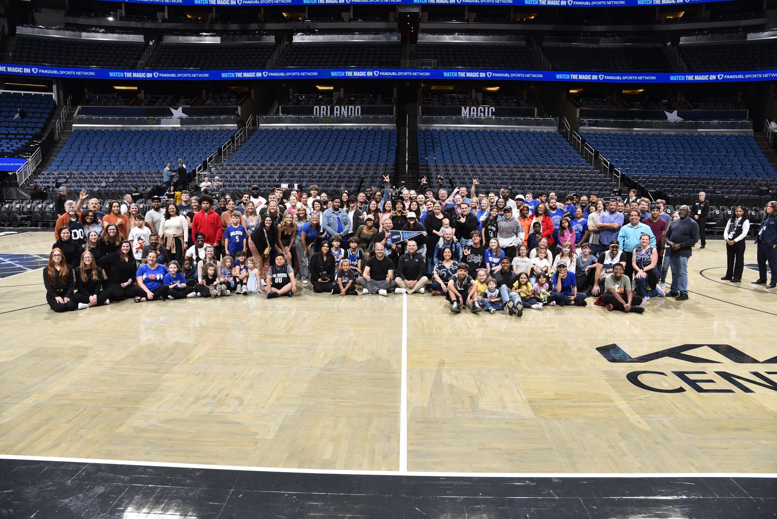 A large group of diverse people, including children and adults, gathered on a basketball court inside an arena, posing for a group photo with empty seats and electronic banners in the background.
