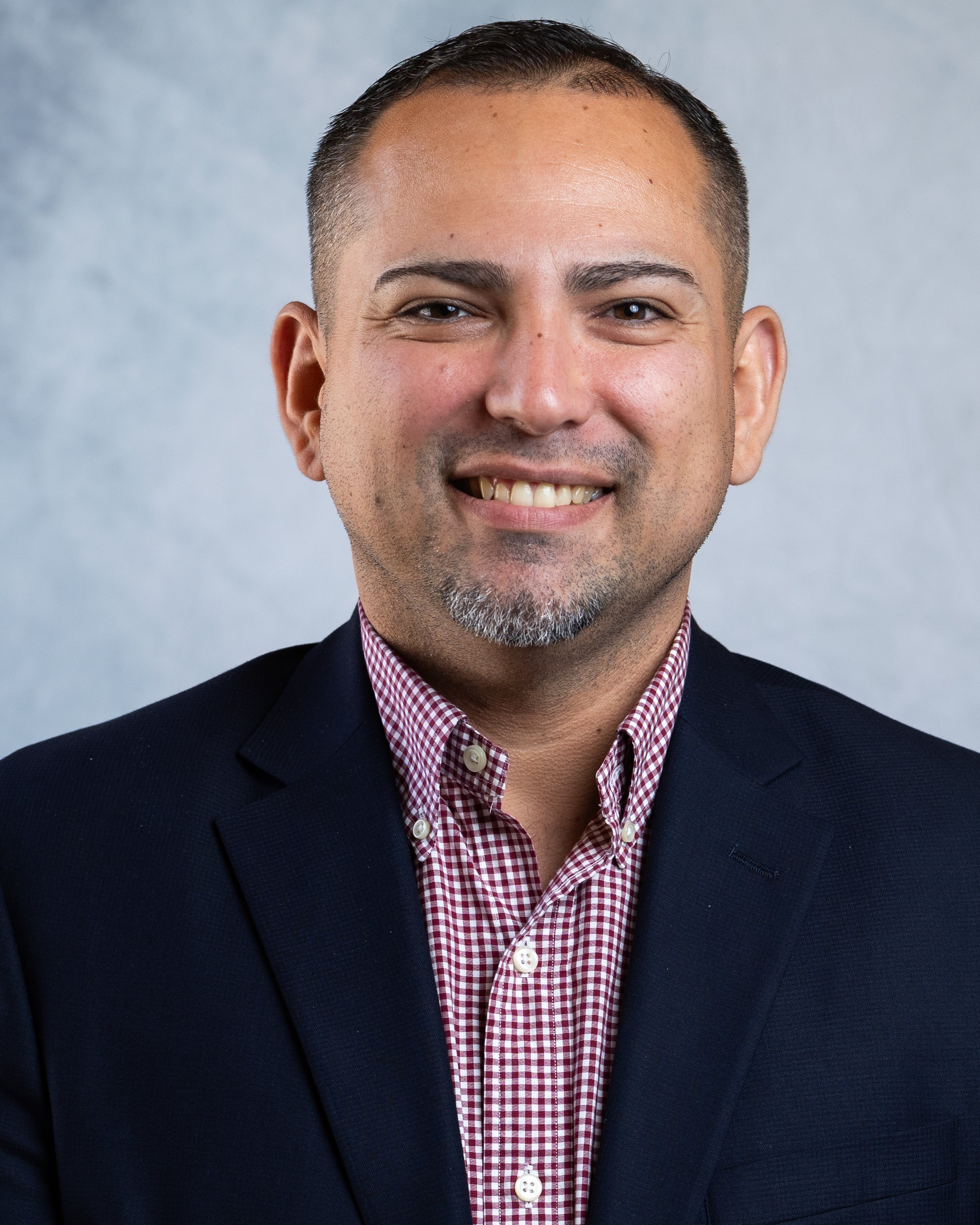 Close-up portrait of a smiling man wearing a dark blazer and a red and white checkered shirt, against a light gray background.