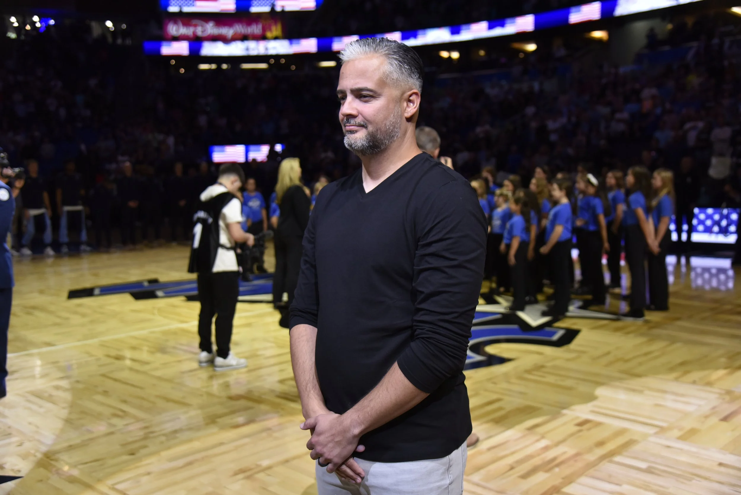 A man with gray hair and beard wearing a black shirt standing on a basketball court with a crowd and team members in the background.