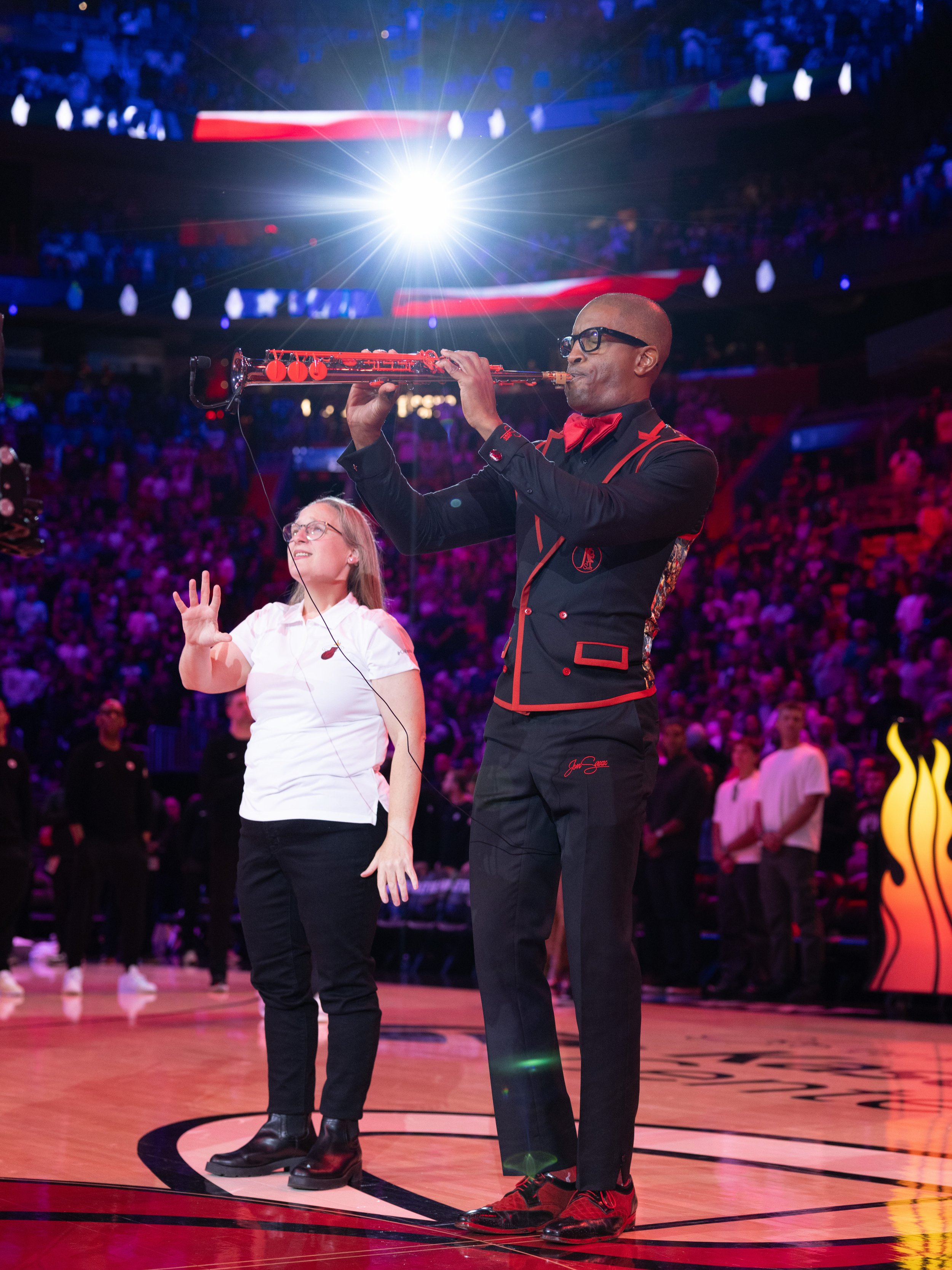 A man playing a saxophone on a basketball court with a woman standing nearby, and a crowd in the background at an event with bright stage lighting.