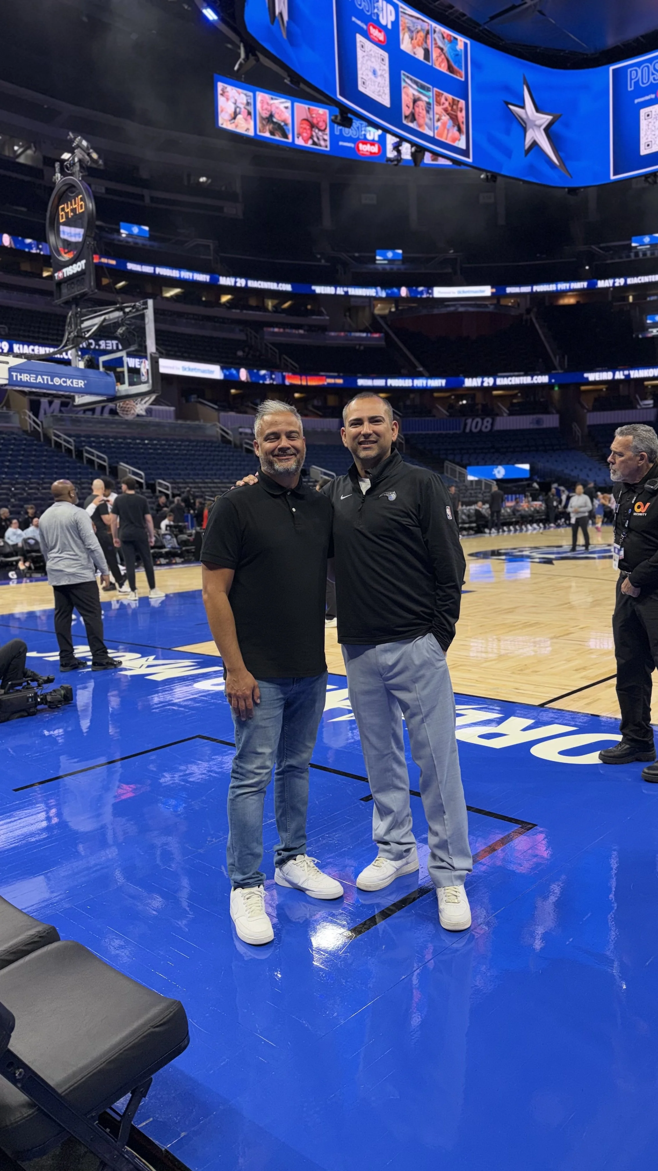 Two men standing on a basketball court inside a sports arena, smiling at the camera. One is wearing a black polo shirt and jeans, the other a black jacket and light-colored pants. There are other people and photographers in the background, with large screens displaying images and information overhead.