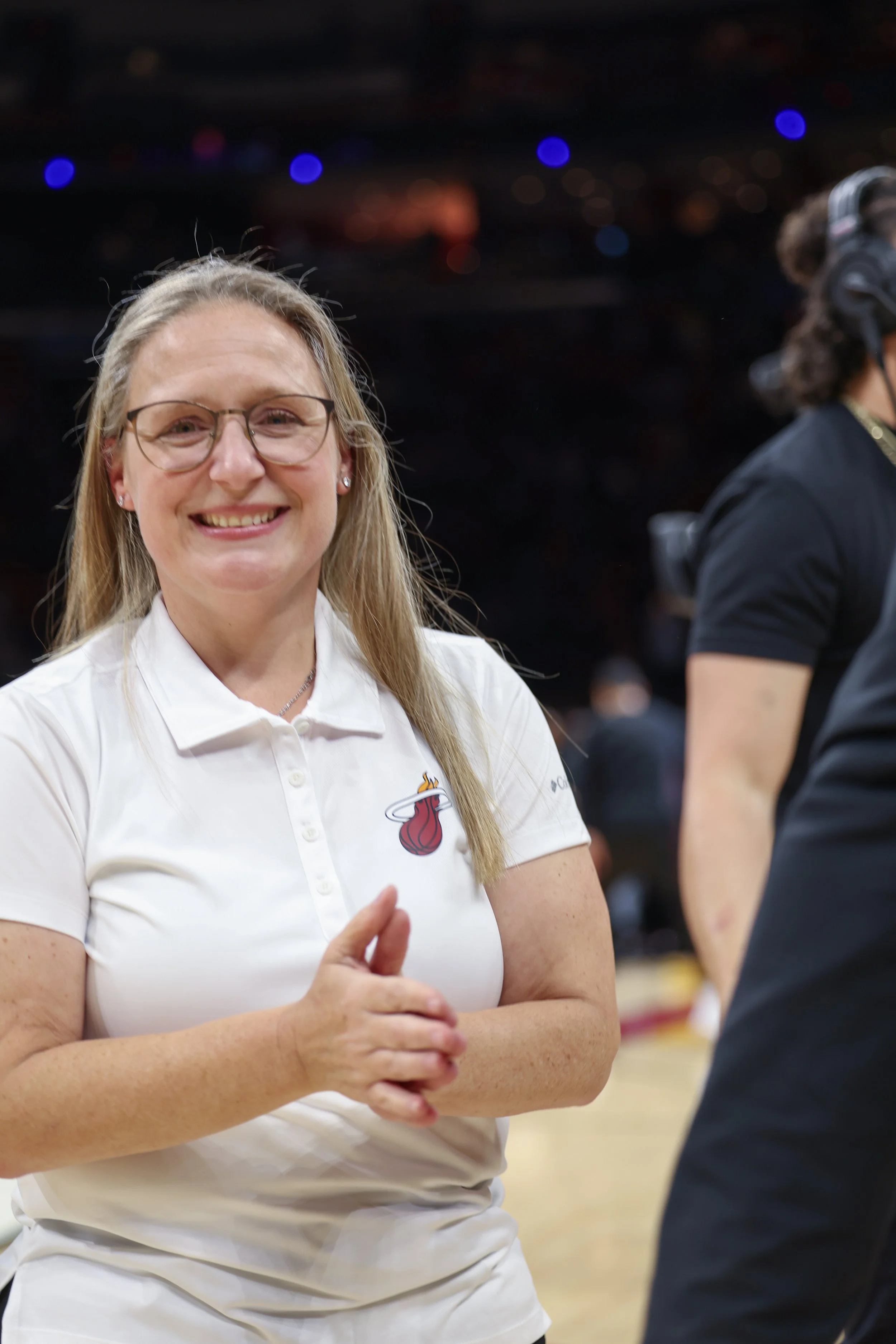 A woman with glasses and long blonde hair smiling, wearing a white polo with a logo on the chest, standing indoors with another person partially visible beside her.